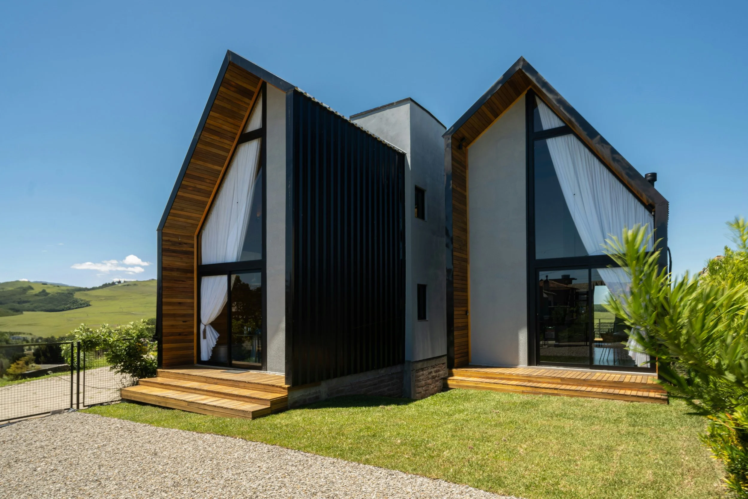 Modern house with steep, asymmetrical rooflines, large windows with white curtains, and a wooden deck, set against green hills and a clear blue sky.
