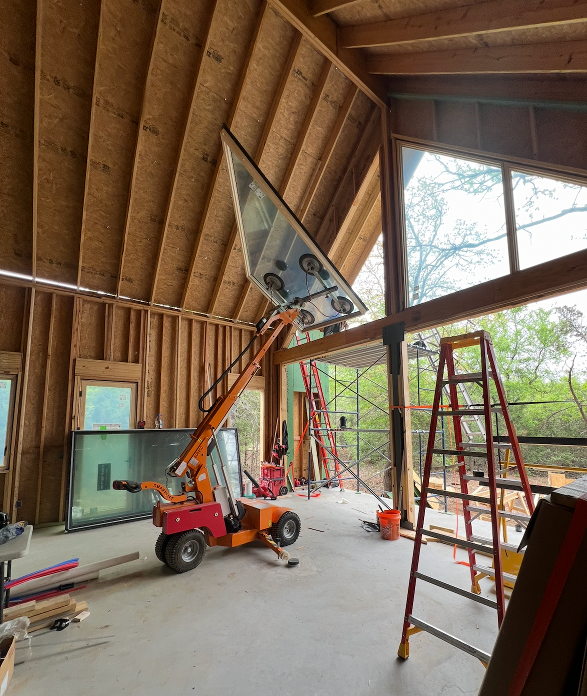 Construction site inside a house with exposed wooden framing and large windows, featuring scaffolding, a ladder, and a crane lifting window panes.