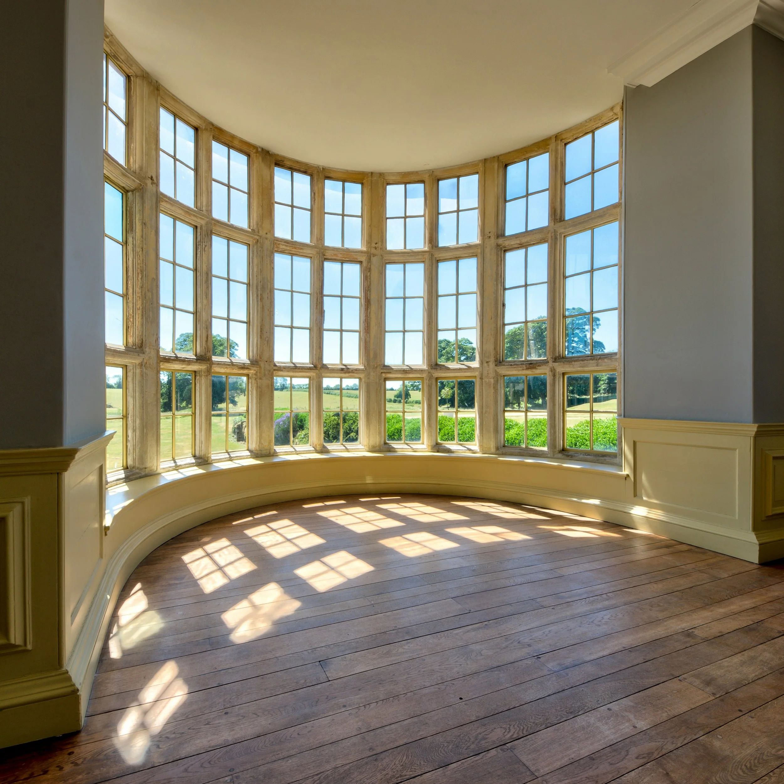A large, curved window with multiple panes, revealing a bright outdoor landscape with green trees and fields, casting shadows on a wooden floor inside.