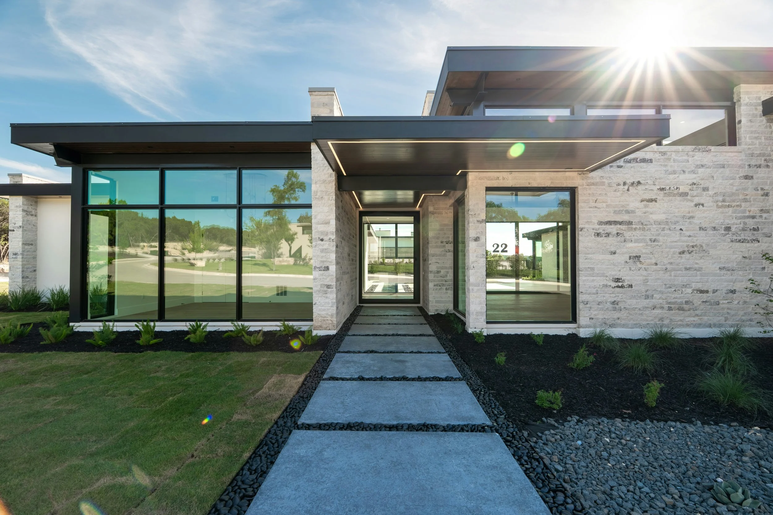 Modern house with large glass windows, a stone exterior, and a pathway leading to the entrance, under a bright sunny sky.