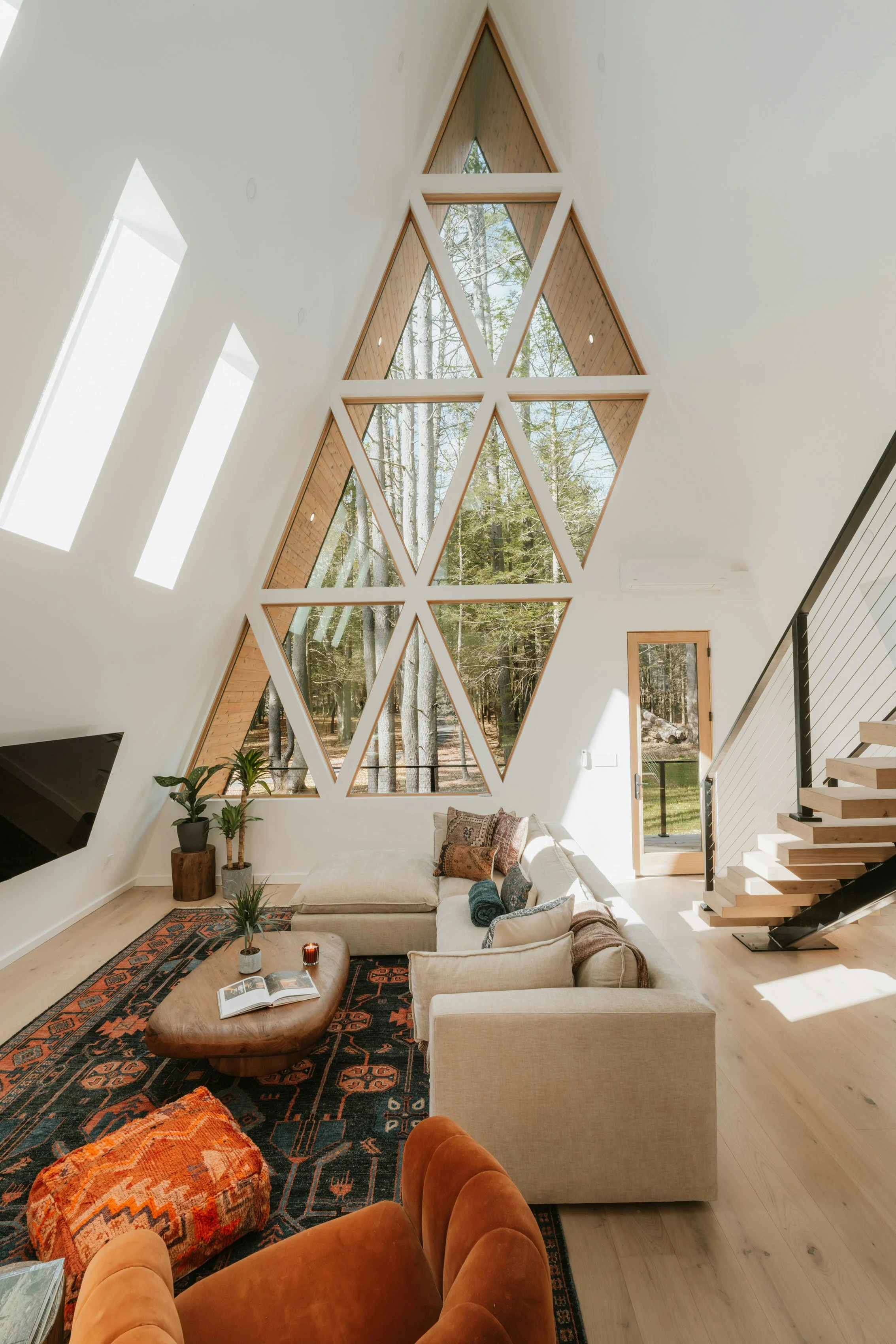 A spacious living room with a high angled ceiling featuring large, geometric triangle windows overlooking a forest. The room has a beige sectional sofa with throw pillows, a wooden coffee table with an open book, a patterned rug, and some potted plants near the wall. There is a staircase with wooden steps on the right side and a door with glass panels leading outside.
