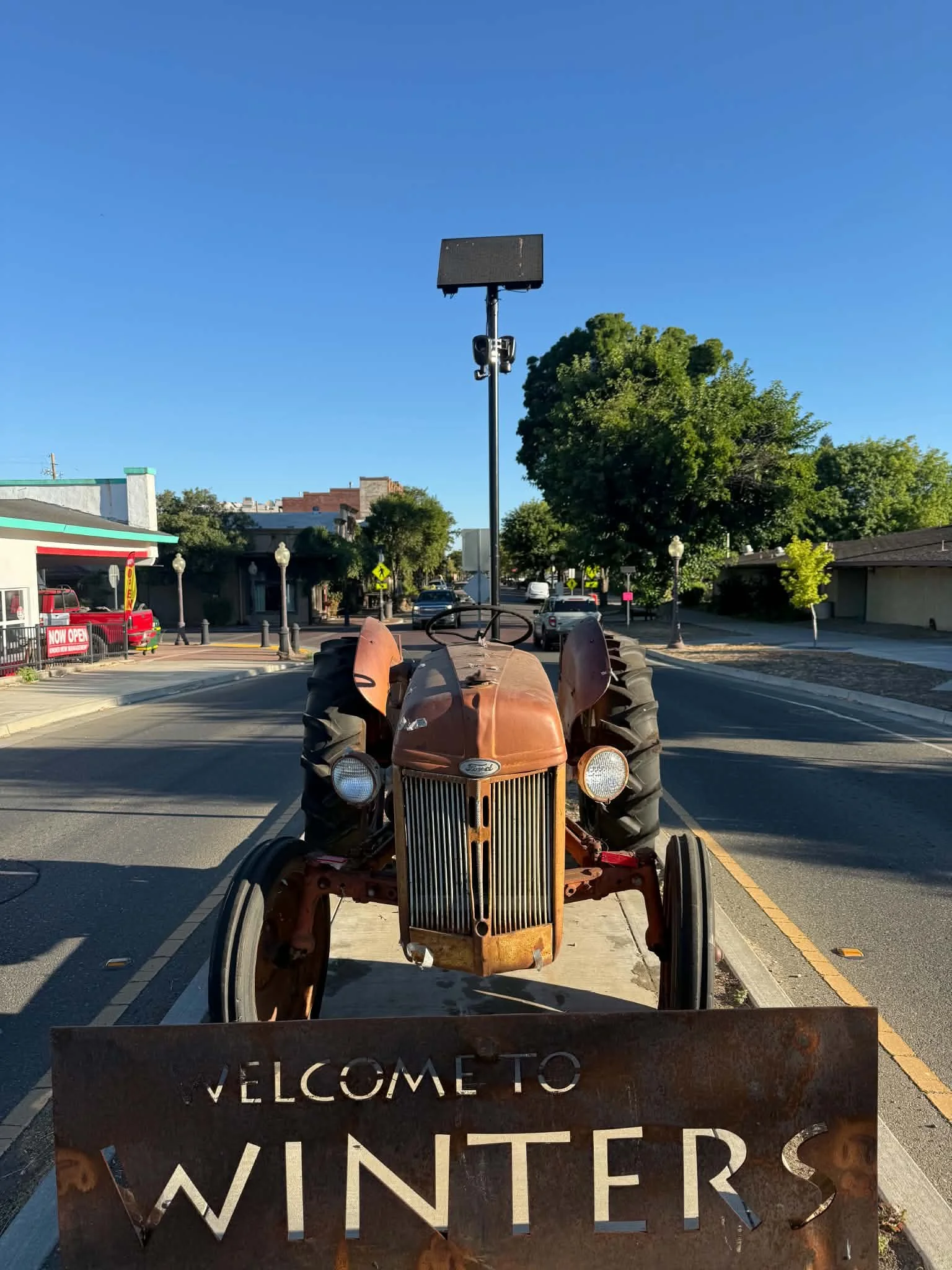 Rusty tractor with a welcome sign in the foreground, a street with cars, trees, and buildings under a clear blue sky in the background.