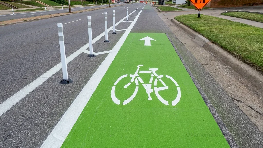 A designated green bike lane with a white bicycle symbol and an upward pointing arrow, separated from the road by white posts.
