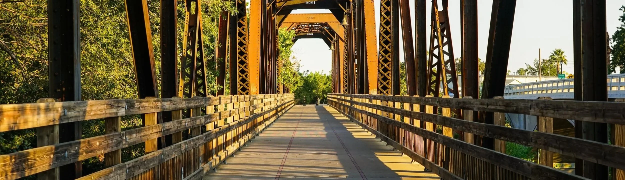 A metal train bridge with a wooden walkway, surrounded by trees with sunlight casting shadows on the walkway.