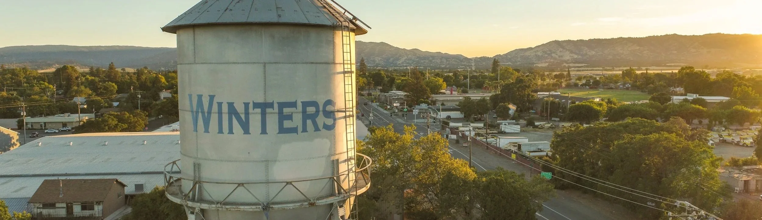 A water tower with the word 'WINTERS' painted on it, overlooking a small town with trees, buildings, and mountains in the background during sunset.