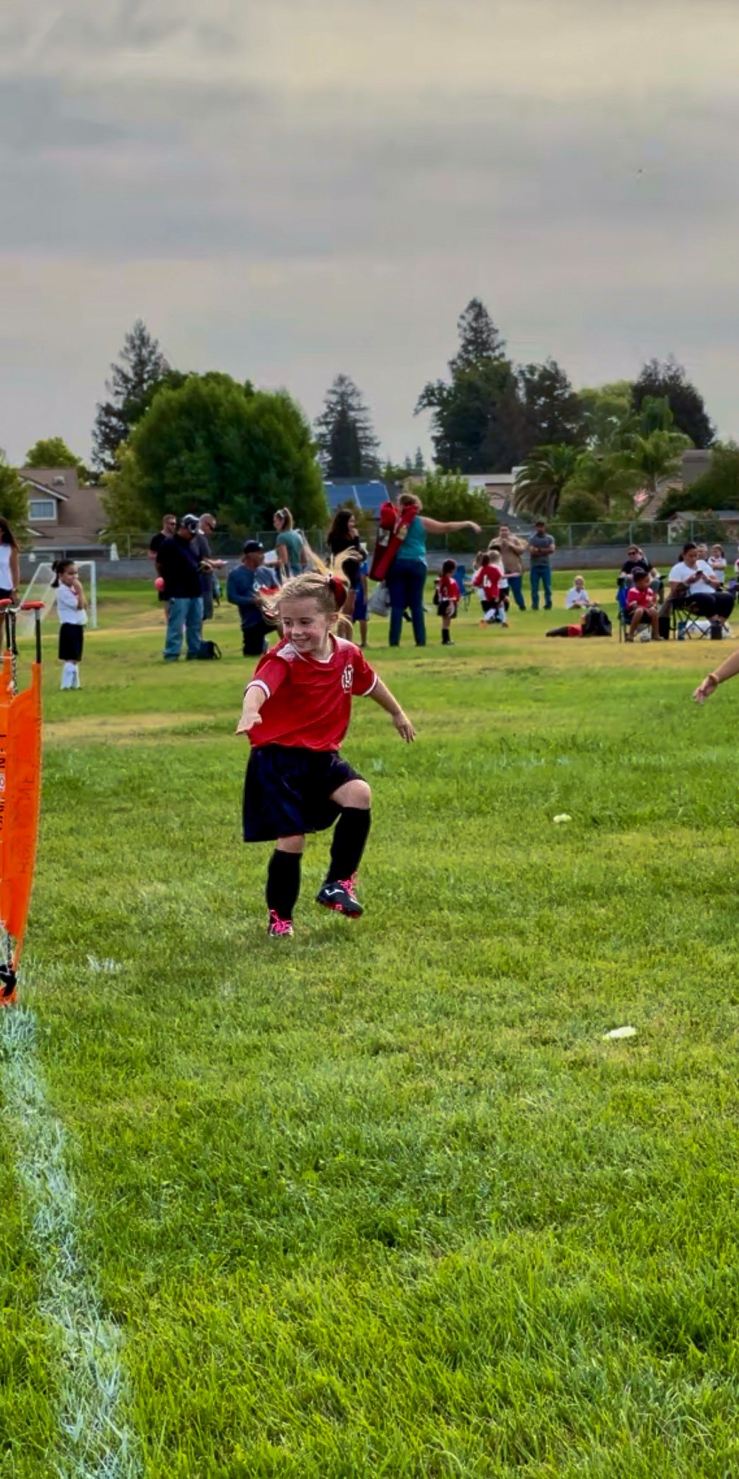 A young girl in a red sports uniform running on a grassy field during a youth soccer game, with spectators and other children in the background. Representing youth sports in Winters CA. Support experienced, competent, effective leadership for Winters