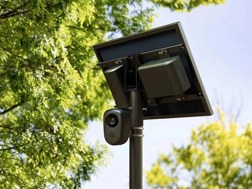 A solar-powered security camera mounted on a pole, with a solar panel attached, outdoors among green trees.