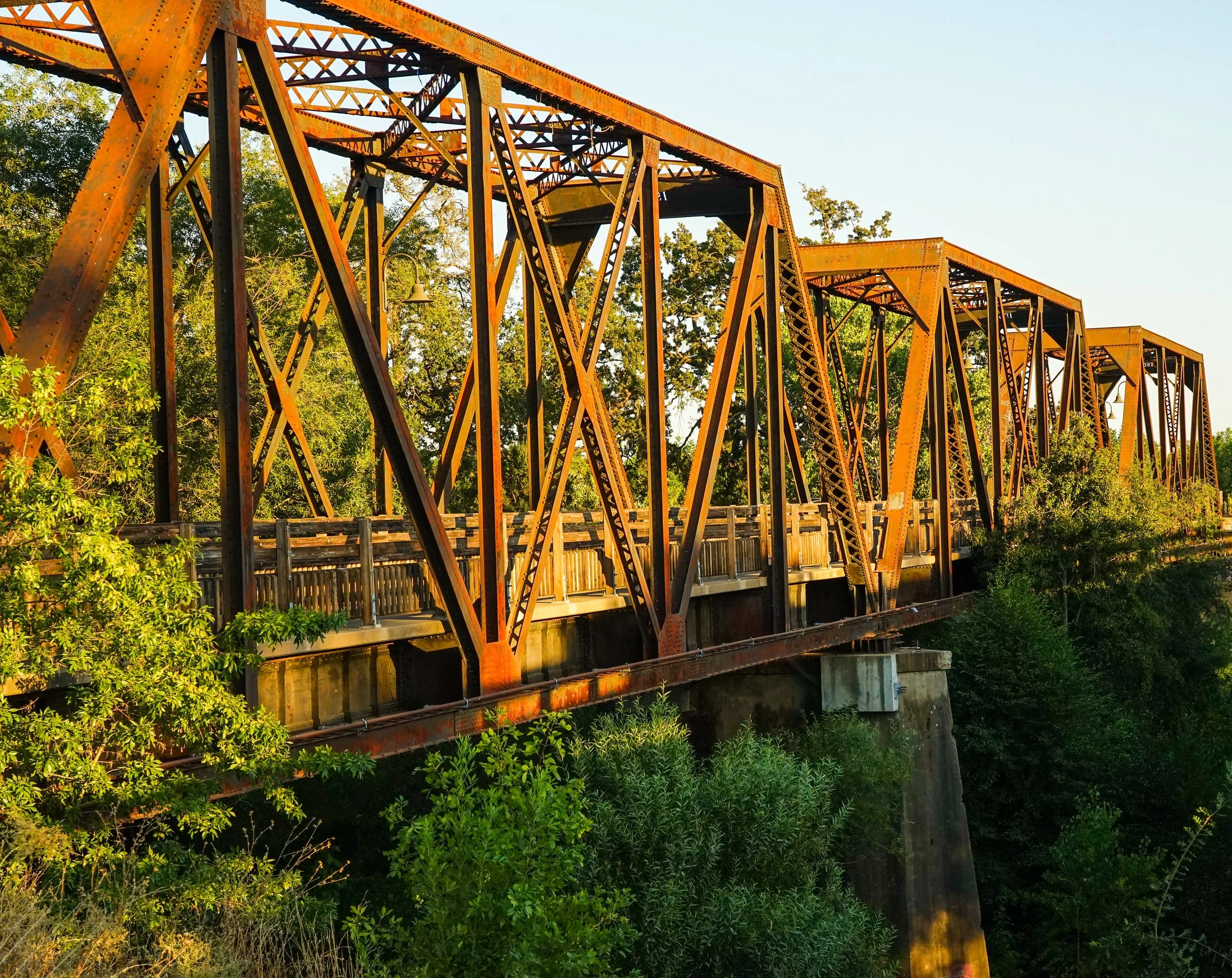 An old rusted metal bridge with a truss structure crossing over a small stream surrounded by green trees and vegetation.