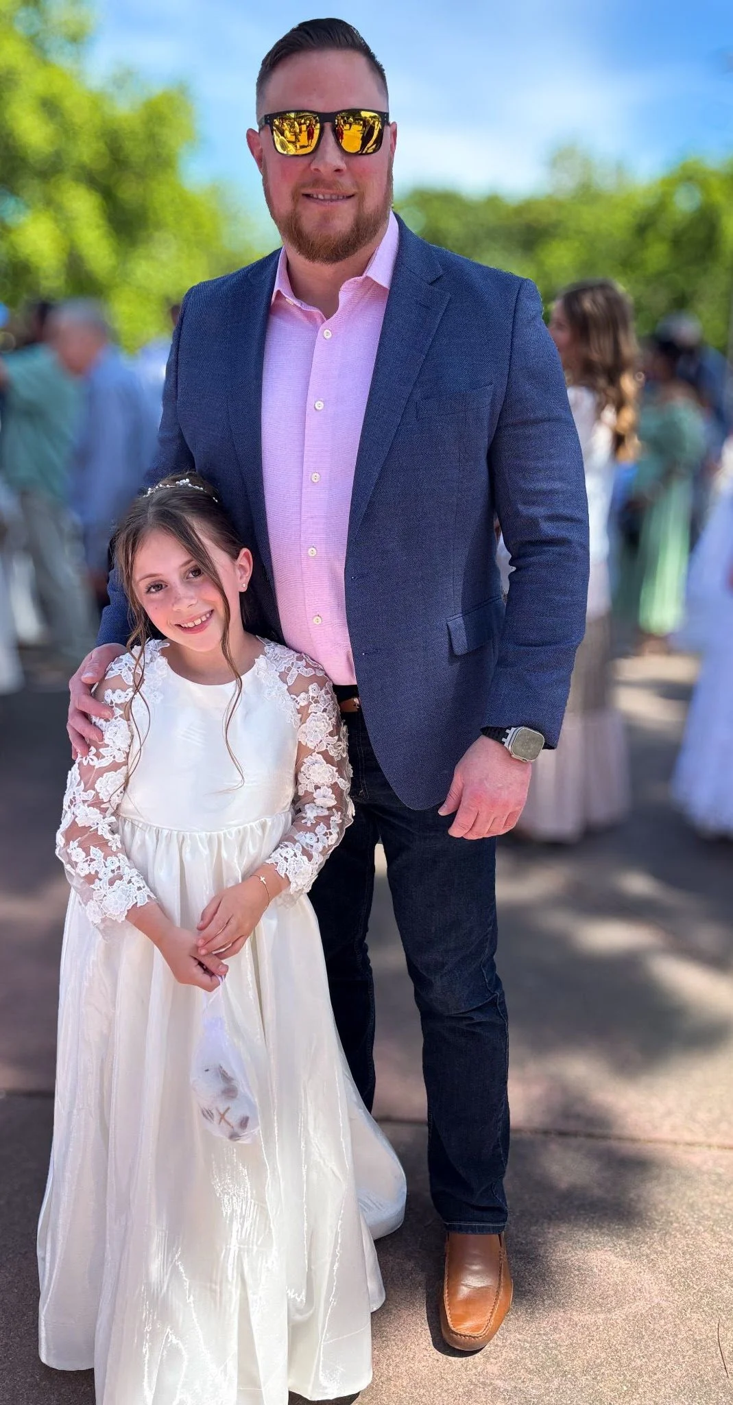 A man and a girl are standing together outdoors at a formal event. The girl is wearing a white dress with lace sleeves, and the man is dressed in a pink shirt, dark blazer, and sunglasses. They are smiling at the camera.