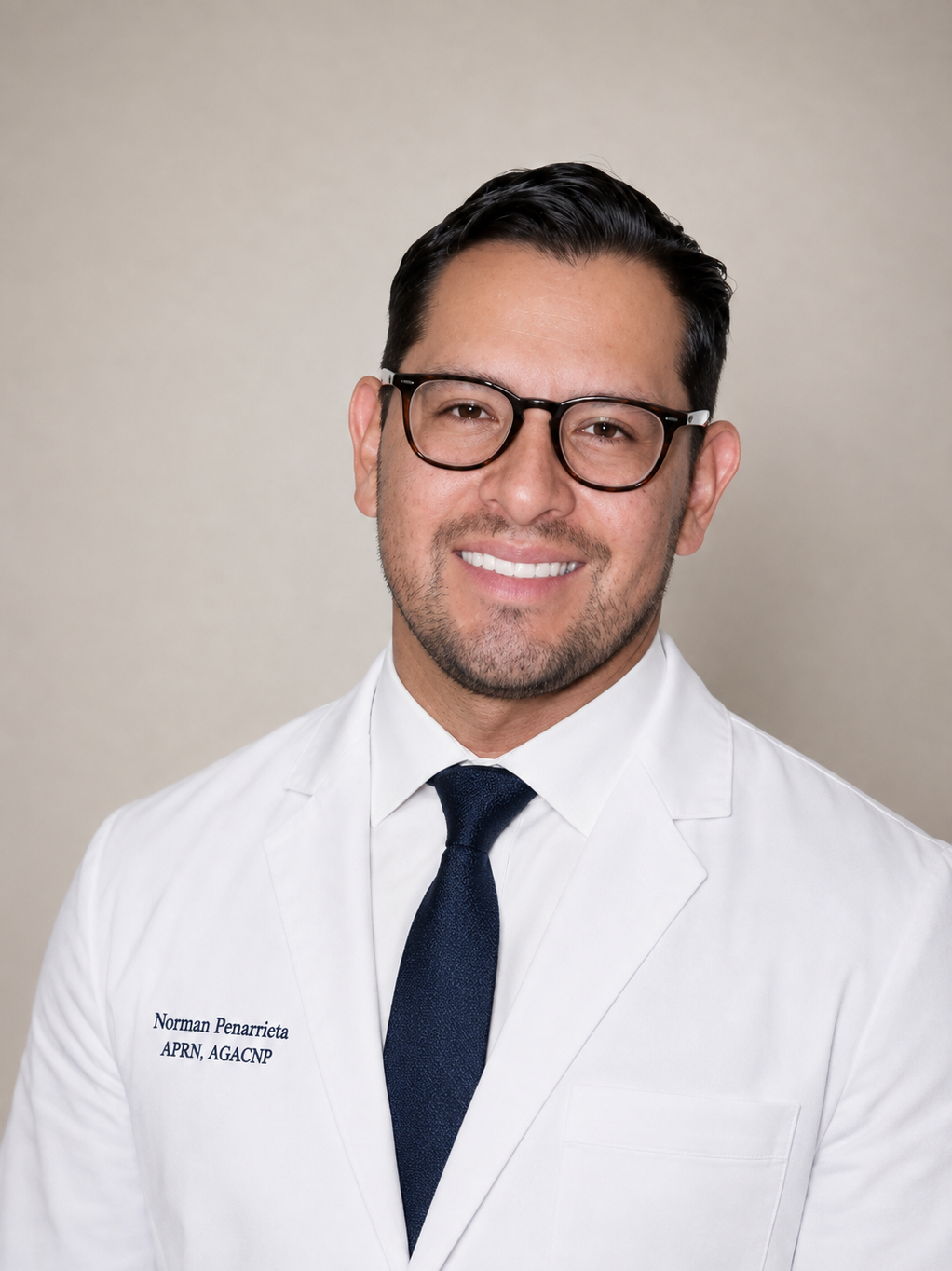 Headshot of a man in a white lab coat with a name tag, wearing glasses, smiling, against a neutral background.