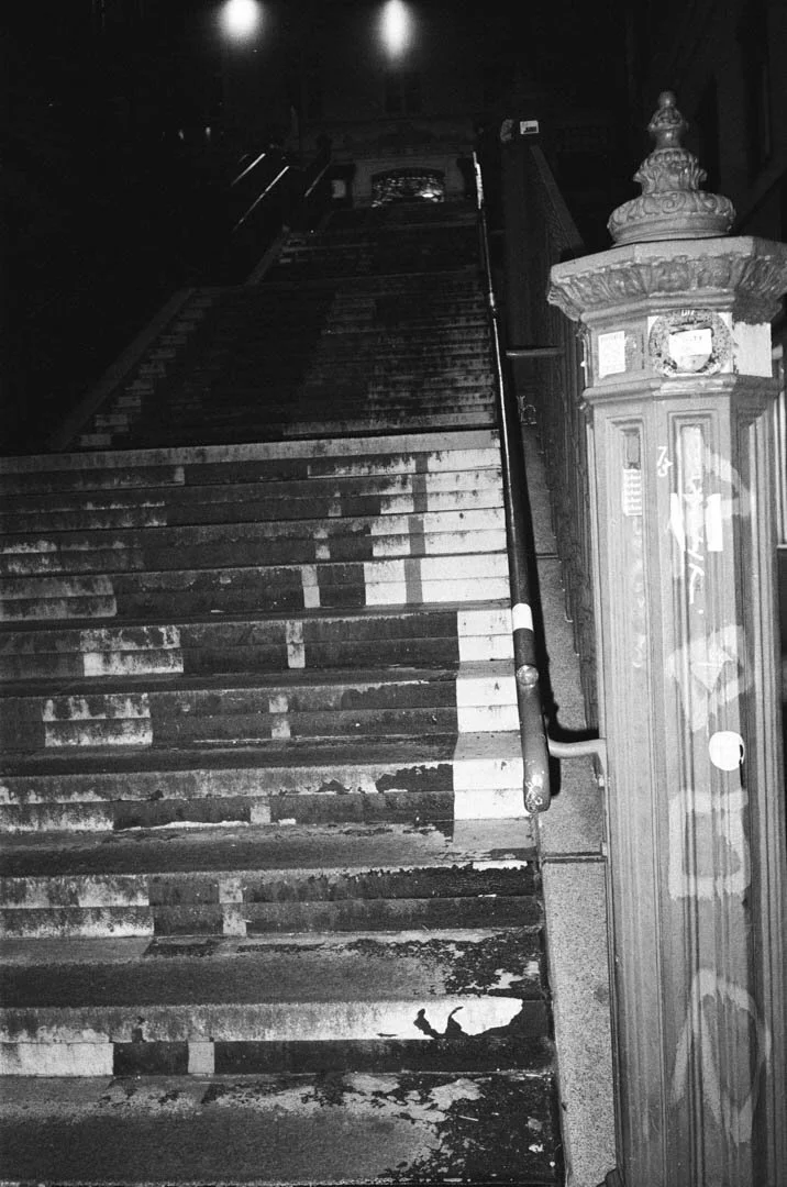 Schmutzige, abgenutzte Treppe mit Handlauf, neben einer Parkuhr, nachts, in Schwarzweiß. Leuchtende Straßenlaternen im Hintergrund.
