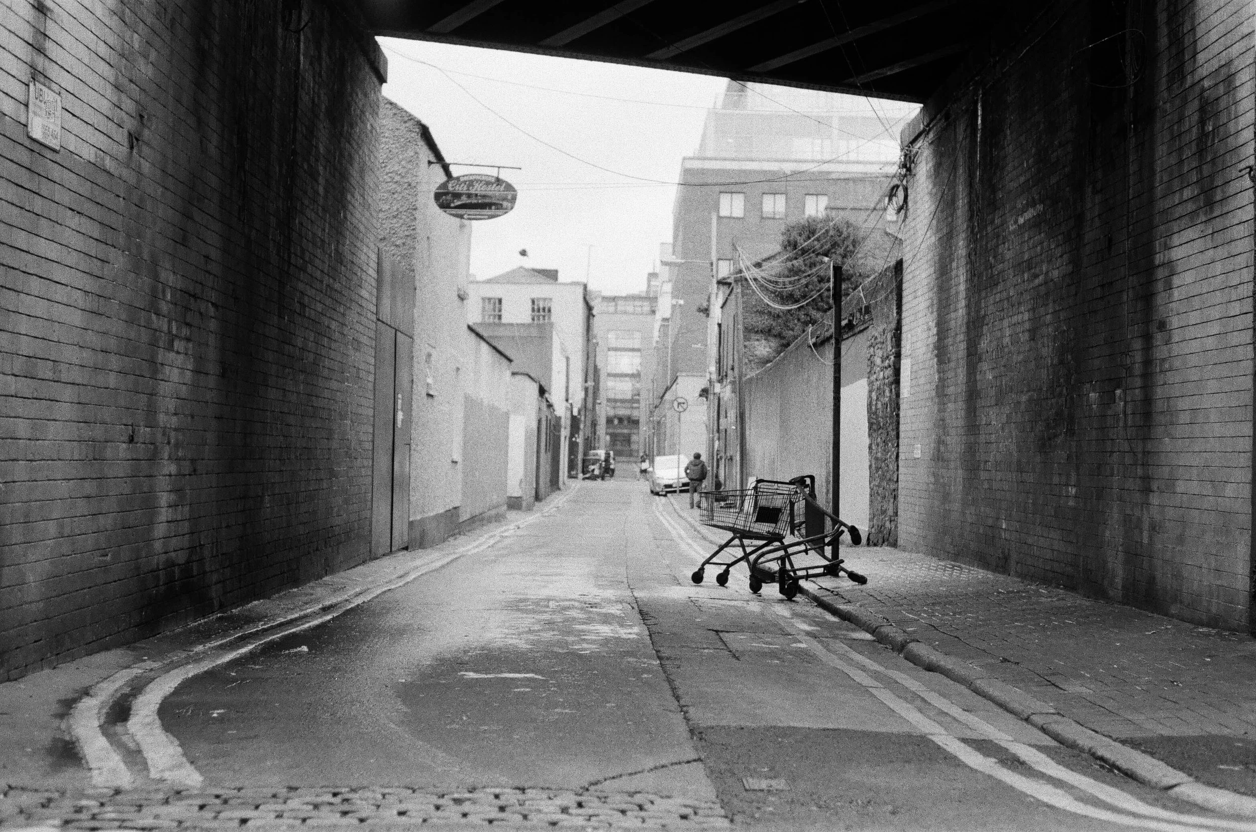 Ein unter einer Brücke gelegener schmaler Stadtweg mit einer Shoppingwagen, der umgekippt auf dem Gehweg liegt, und einer Person in der Ferne, die den Weg entlangläuft.