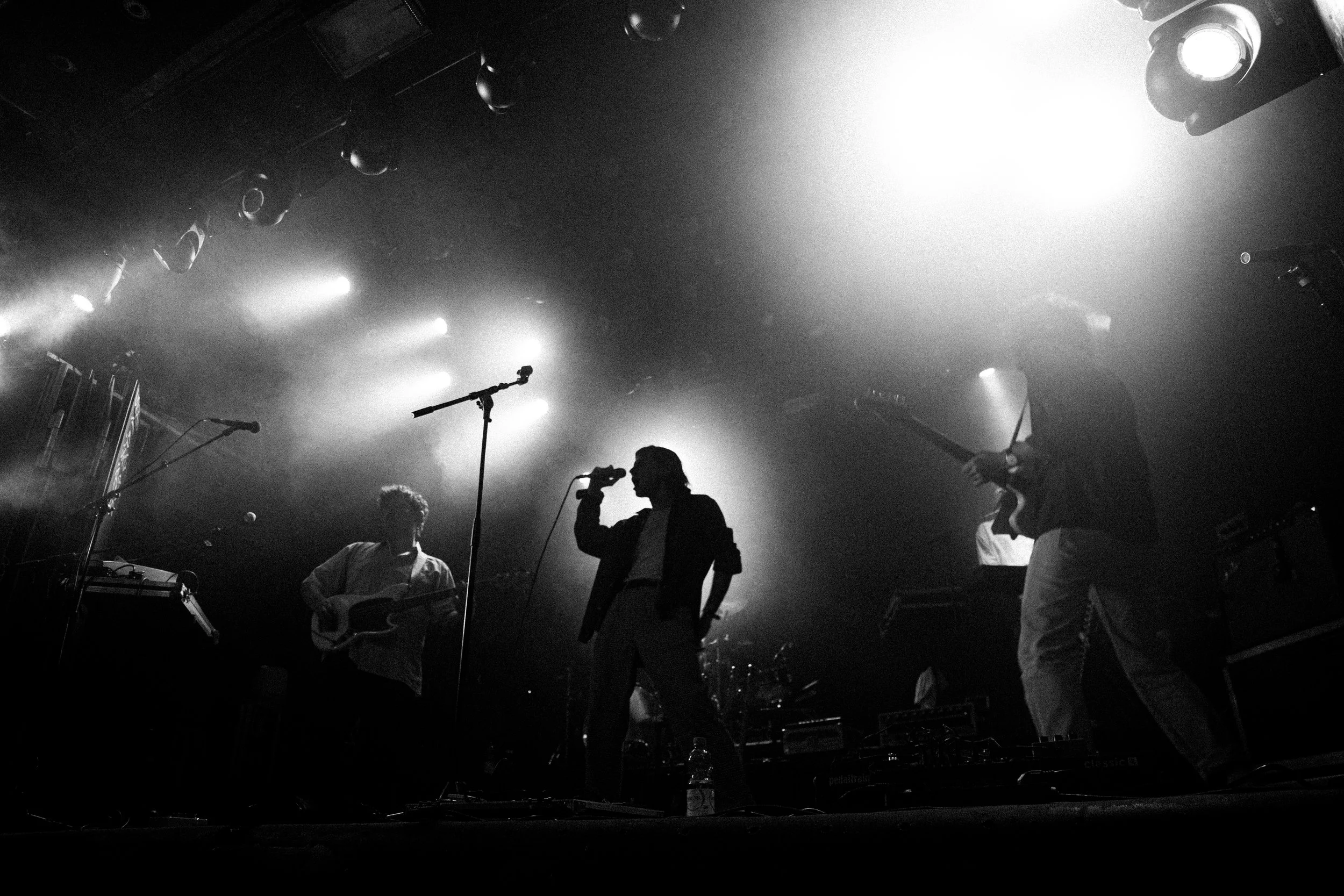 Silhouetten von Musikern auf einer Bühne unter Scheinwerfern, eine Person singt in ein Mikrofon, während andere Instrumente spielen.