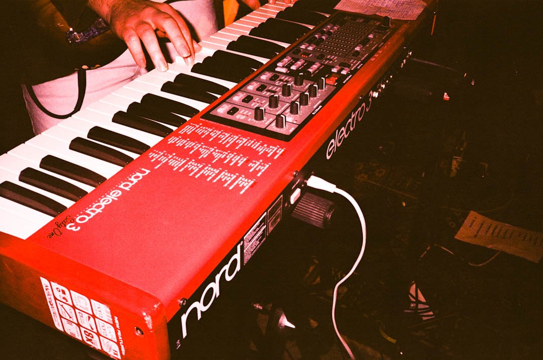 Ein rotes Nord Electro 3 Klavier mit schwarzen und weißen Tasten, auf einem Holzständer. Ein Mensch spielt darauf. Das Klavier ist mit Kabeln verbunden.