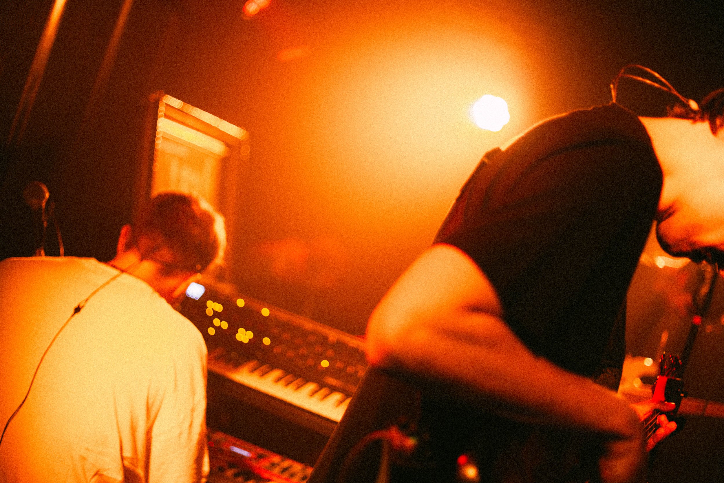 Menschen bei einem Musikproduktions- oder DJ-Event, mit einem Keyboard und einem Laptop, in einer dunklen, orange beleuchteten Umgebung.