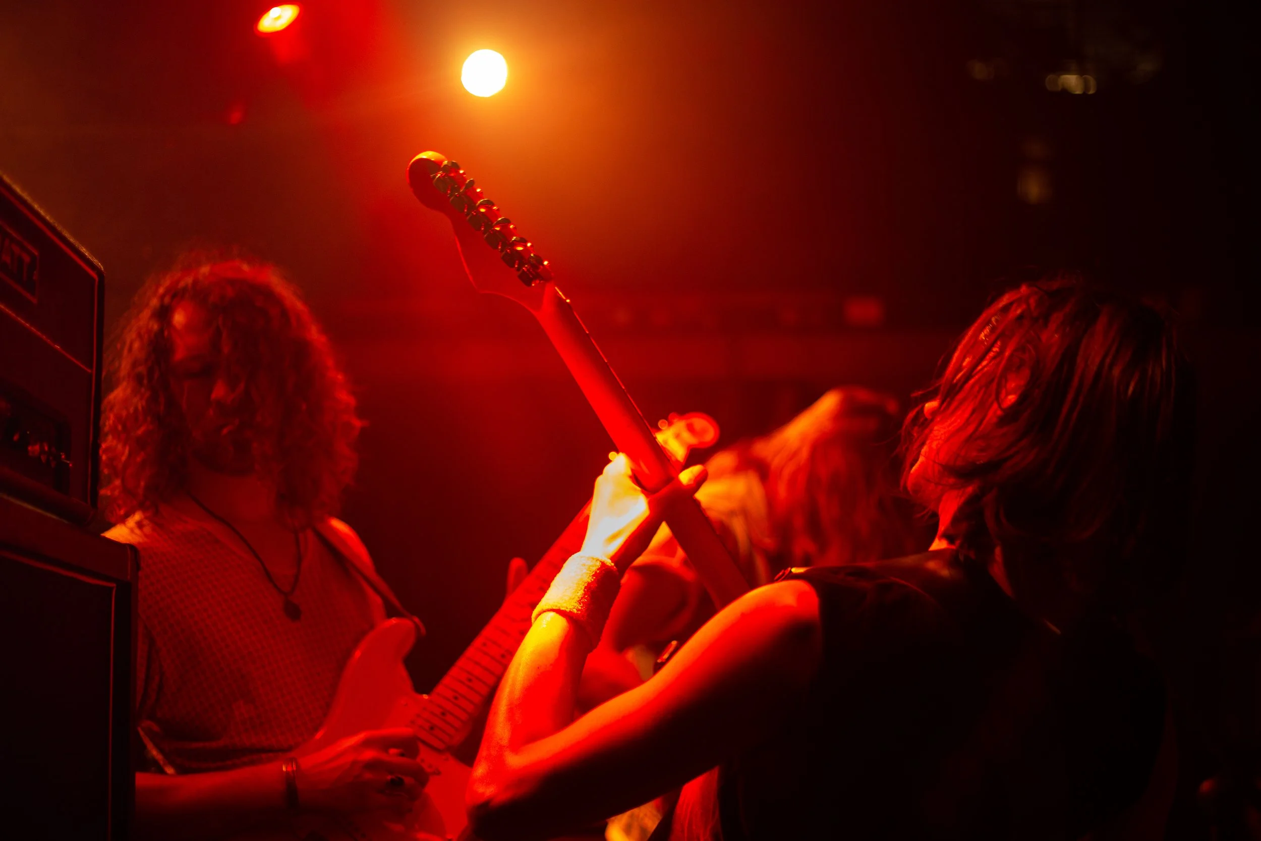 Menschen bei einem Konzert, rot beleuchtet, eine Person hält eine E-Gitarre hoch, andere Personen mit gewundenen Haaren sind zu sehen.