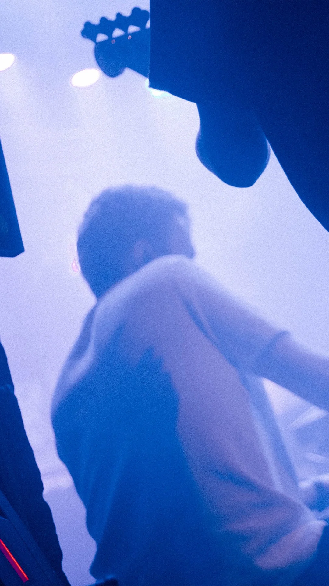 Ein junger Mann mit dunklem Haar trägt eine weiße Jacke und steht in einem Raum mit bläulichem Licht. Die Aufnahme erfolgt von unten nach oben, sodass nur die Silhouette seines Kopfes und Oberkörpers sichtbar sind.