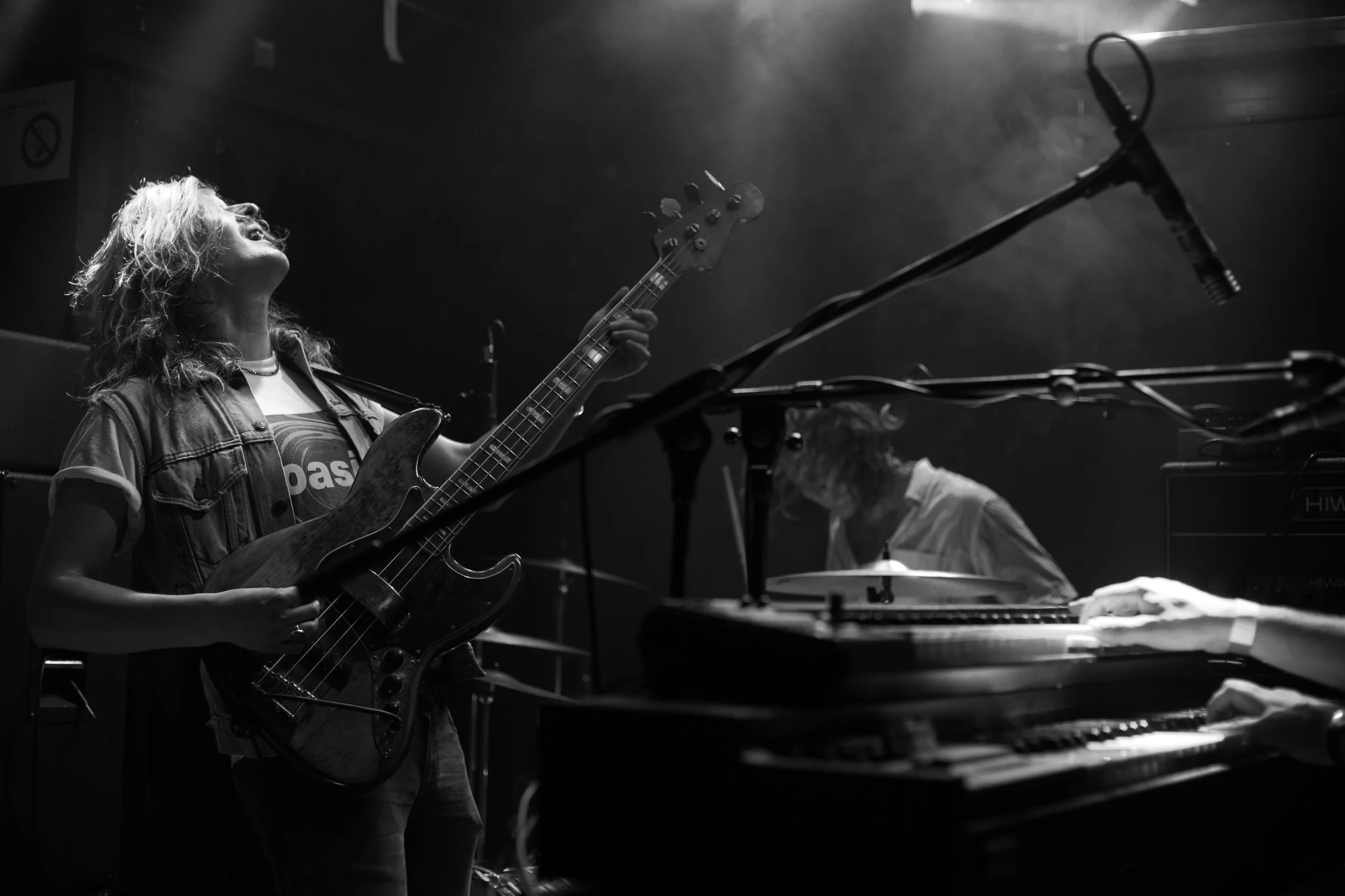 Eine Frau spielt E-Gitarre auf einer Bühne bei einem Auftritt, im Hintergrund ist ein another Musiker am Klavier sichtbar, die Szene ist schwarz-weiß gehalten.