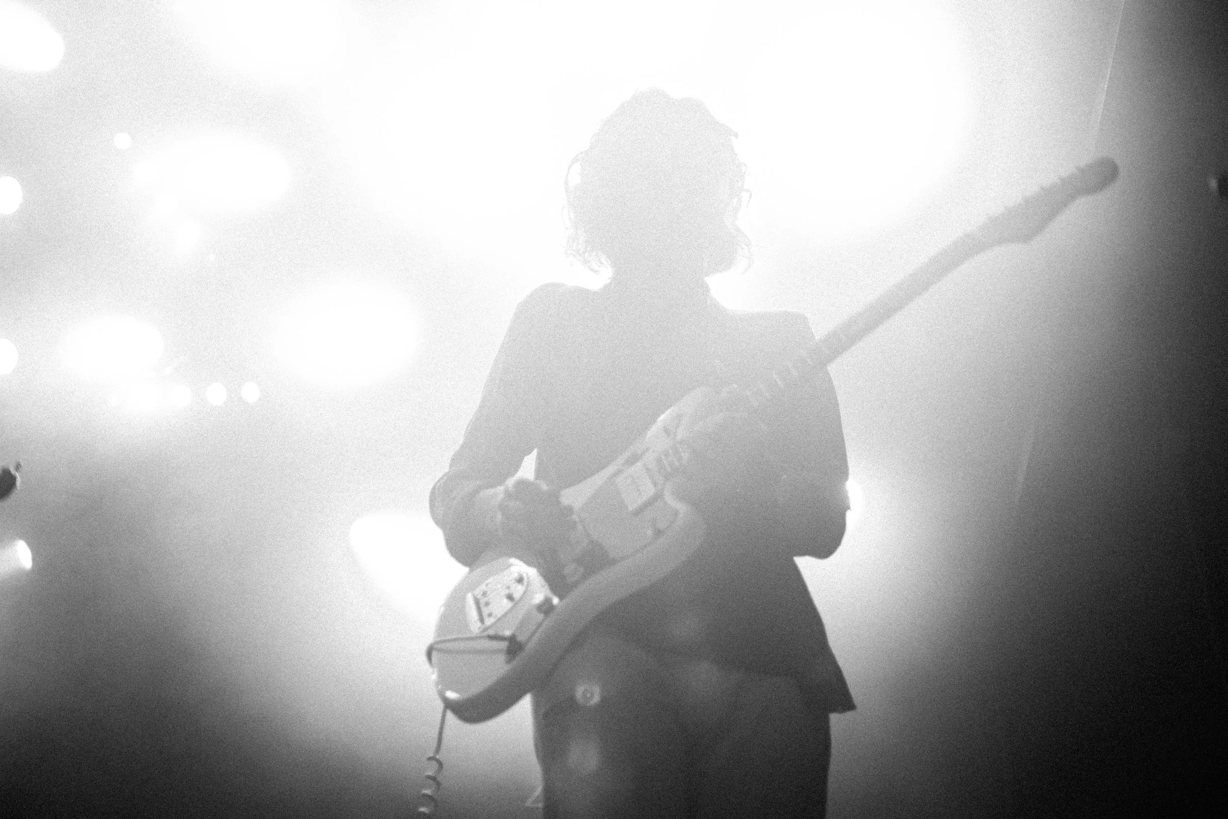 Schwarzweißfoto eines Gitarrenspielers auf einer Bühne mit hellem Licht im Hintergrund.