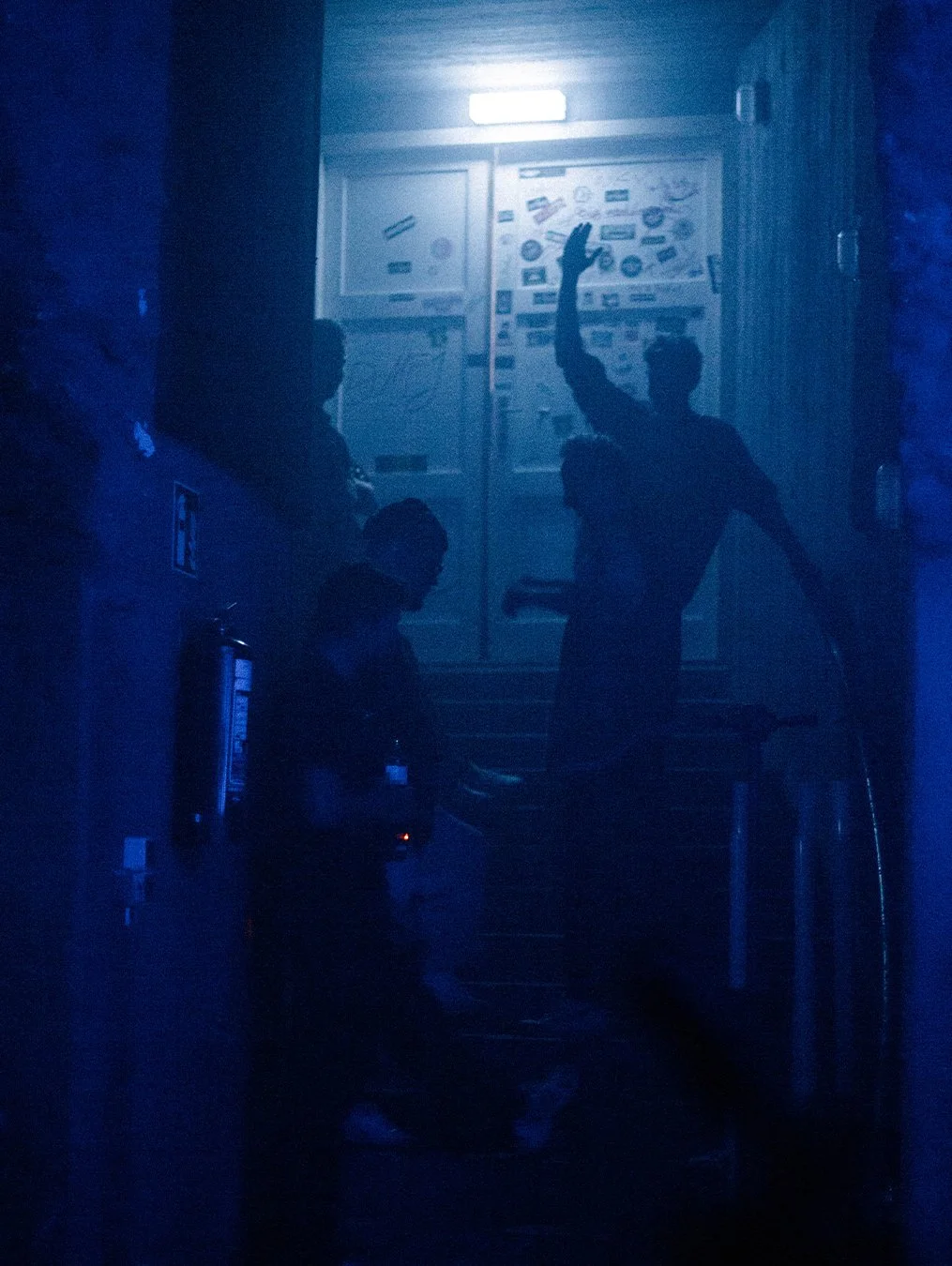 Drei Personen in einem dunklen, bläulichen Raum im Gespräch, eine Person hebt die Hand. Im Hintergrund sind Poster und ein Licht zu sehen.