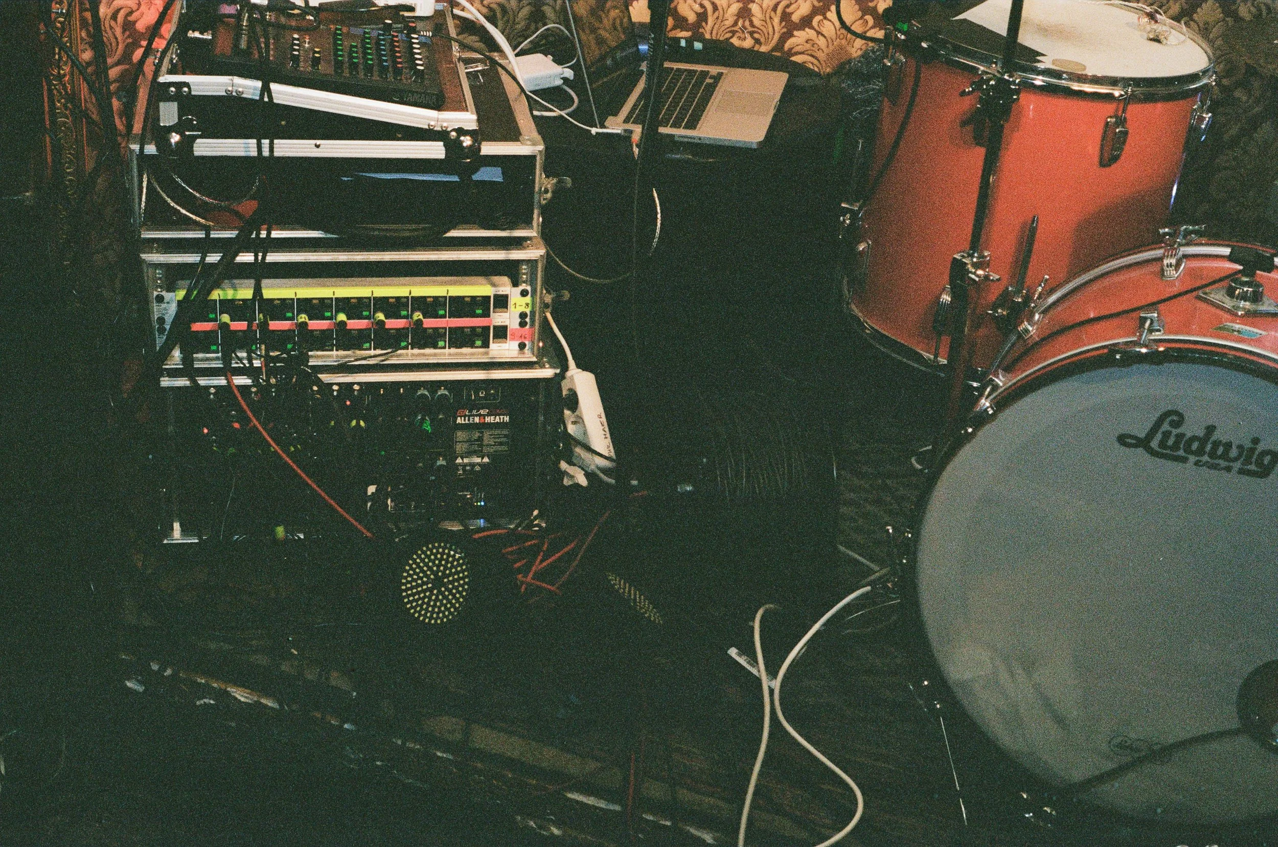 Musikinstrumente und technische Geräte, darunter ein Schlagzeug, eine Laptop, ein Mischpult und Kabel, in einem Raum mit Wandtapete.