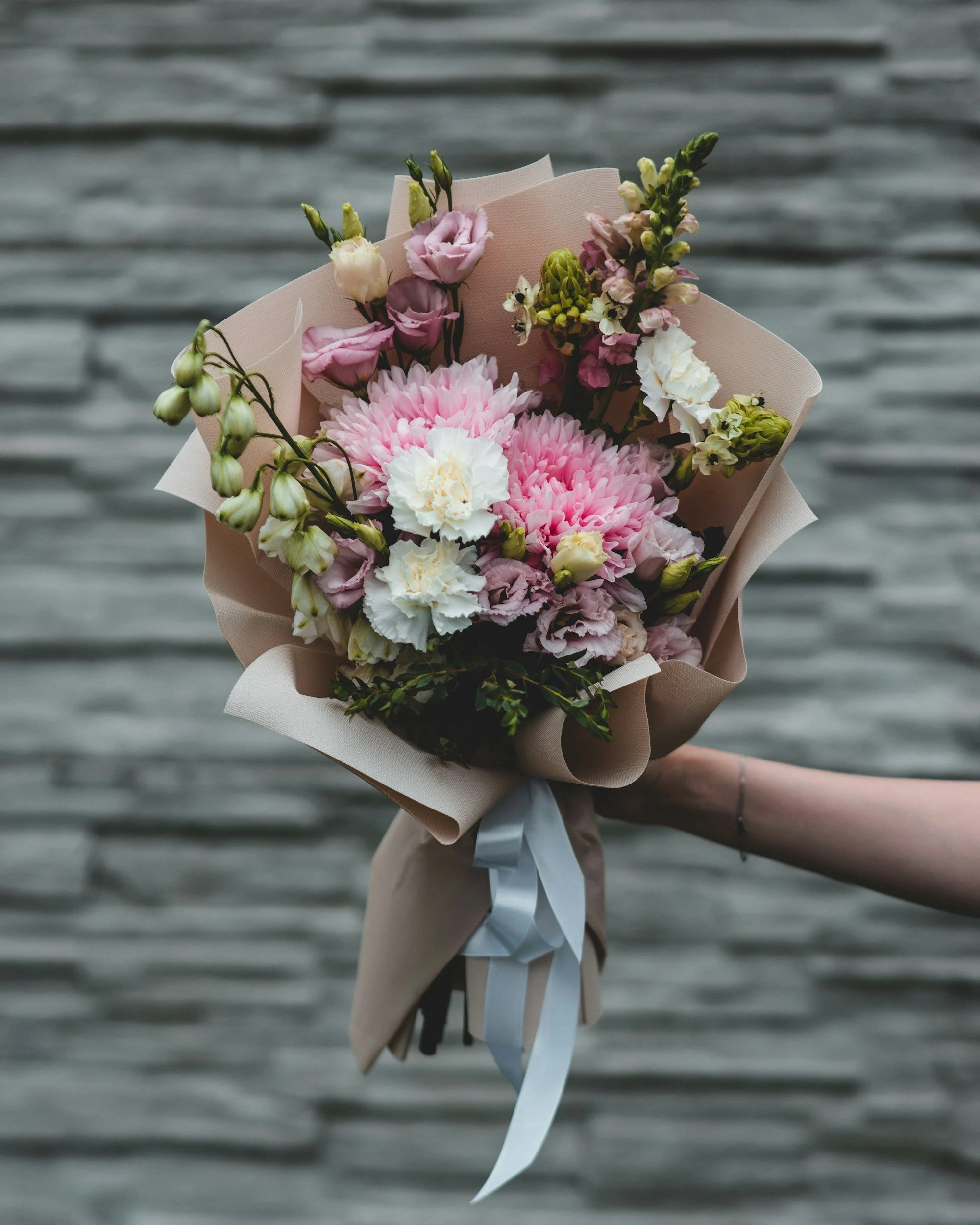 A hand holding a bouquet of pink and white flowers wrapped in beige paper against a background of wooden planks.