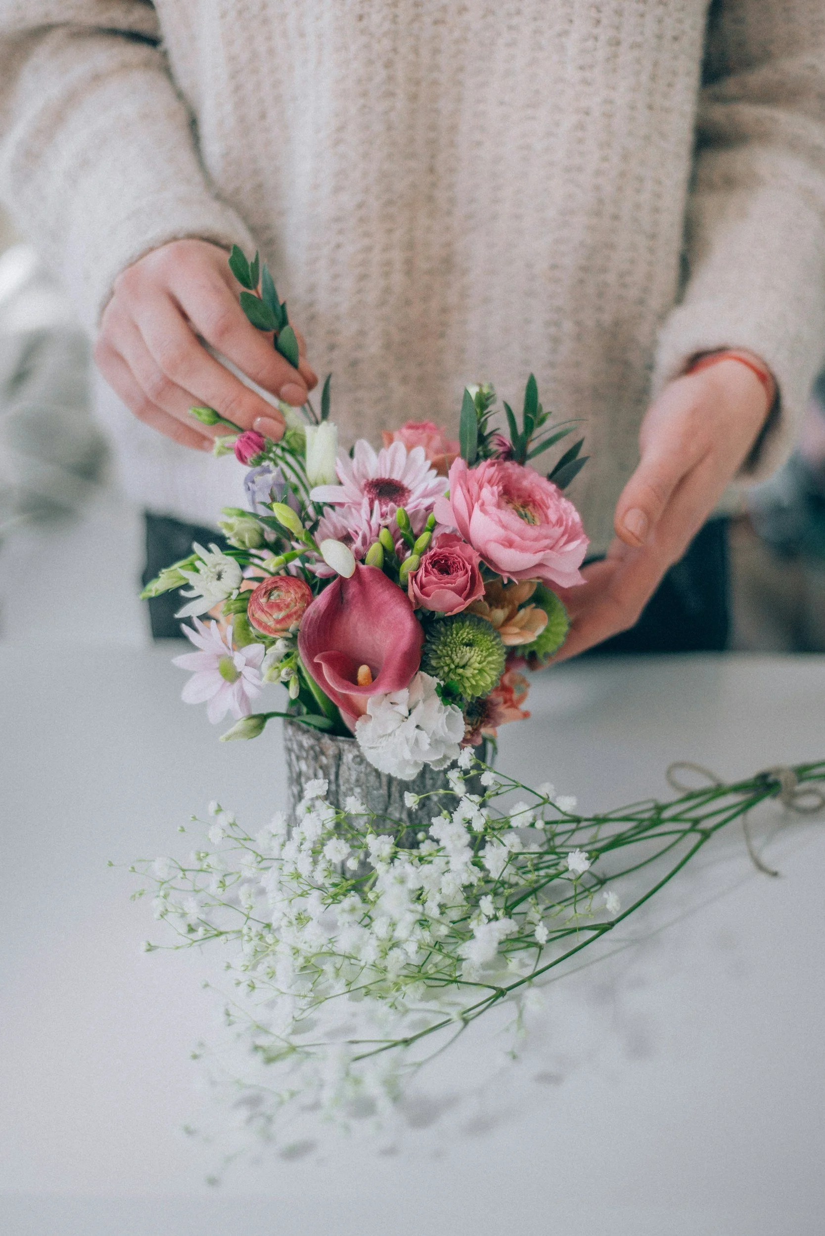 A person arranging a bouquet of pink, white, and purple flowers in a silver vase on a white surface. A bunch of white baby's breath flowers is placed on the table in front.