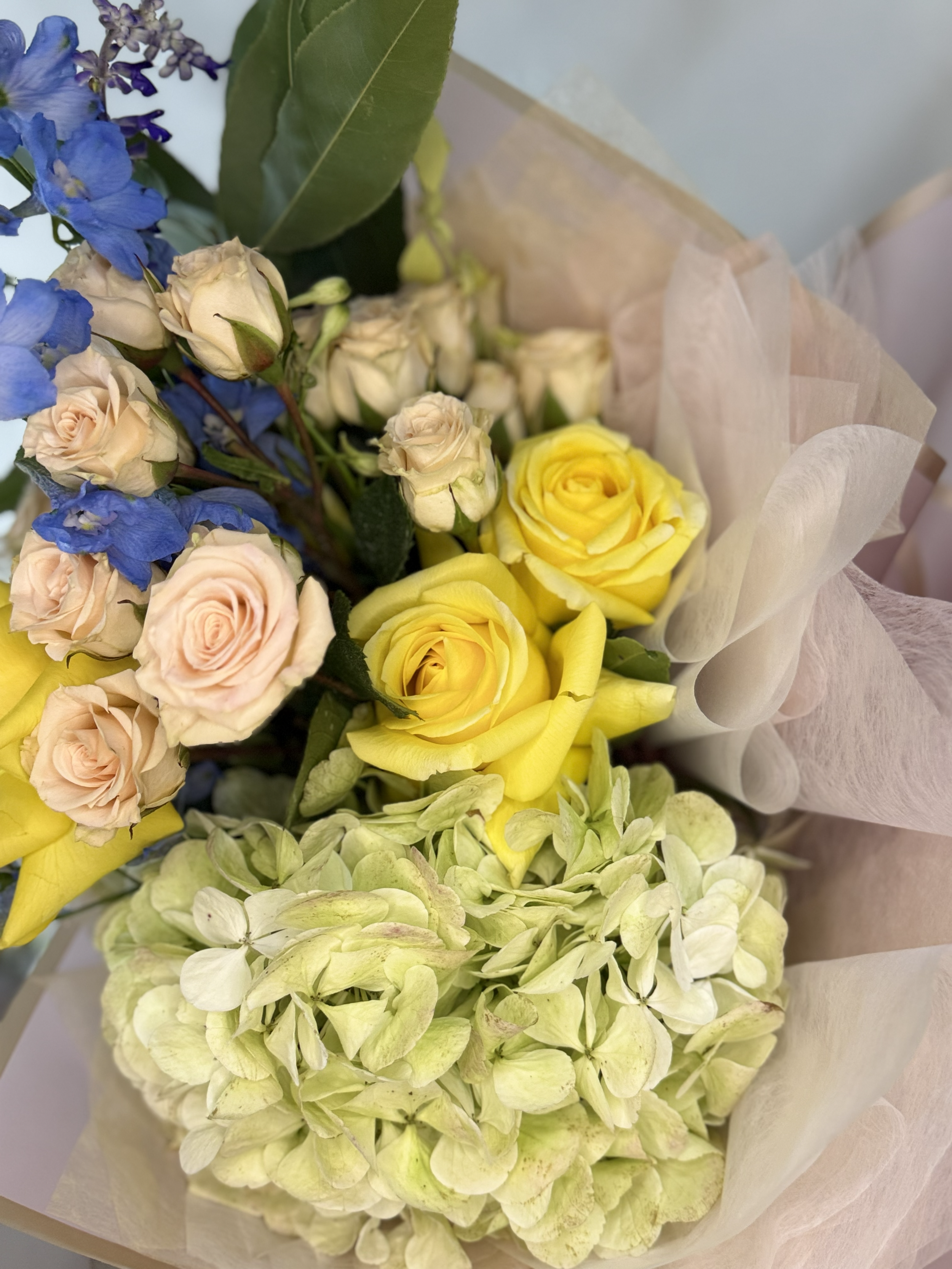 Bouquet of peach and yellow roses, blue delphiniums, white hydrangeas, and greenery wrapped in beige paper.