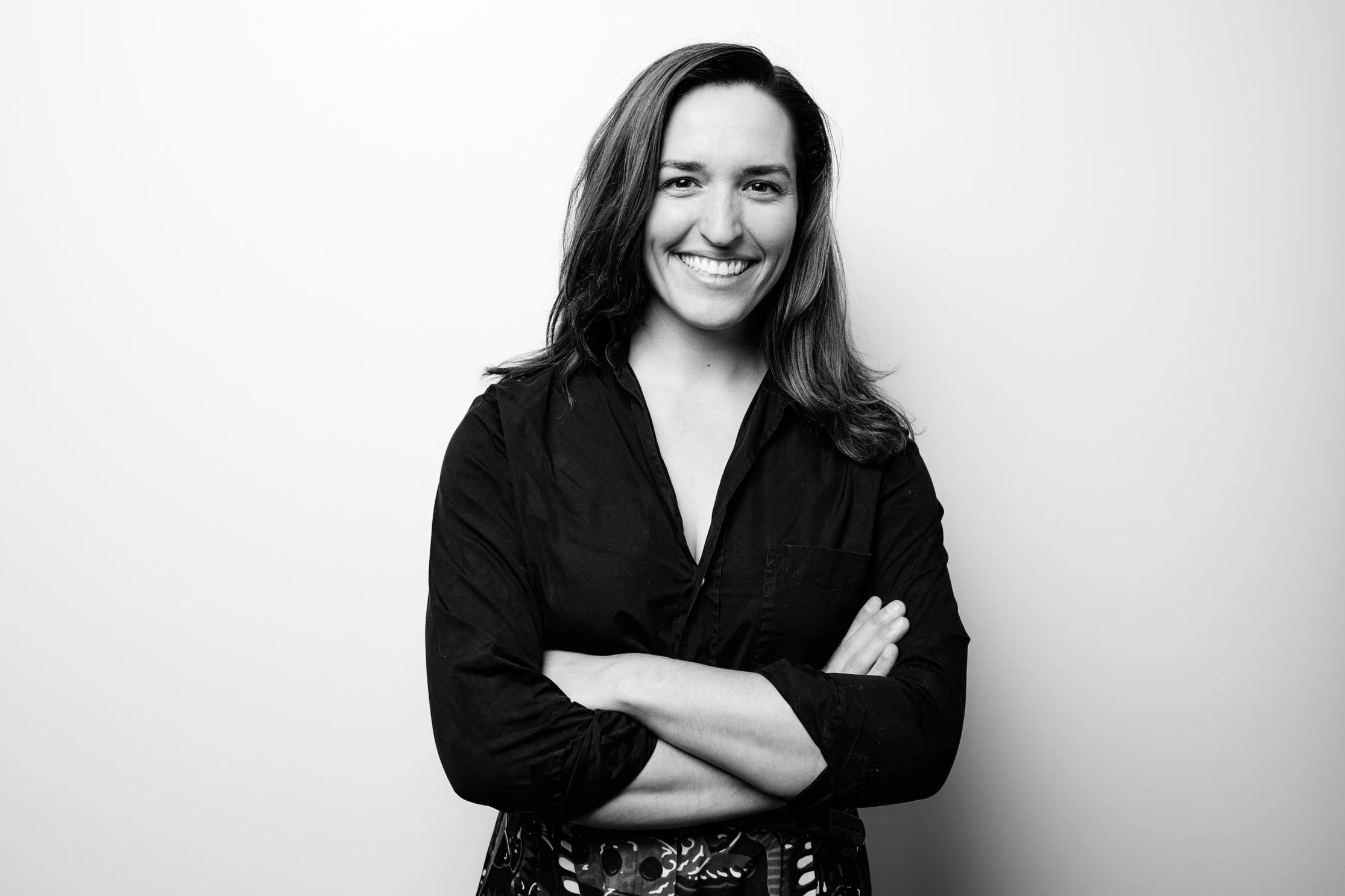 Black and white photo of a woman smiling with arms crossed, wearing a black shirt and patterned pants, standing against a plain background.