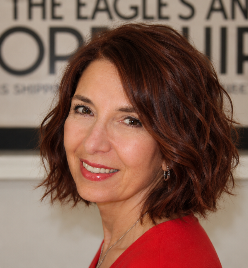 A woman with short, wavy auburn hair, wearing earrings and a red top, smiling at the camera in front of a sign that has partially visible text.