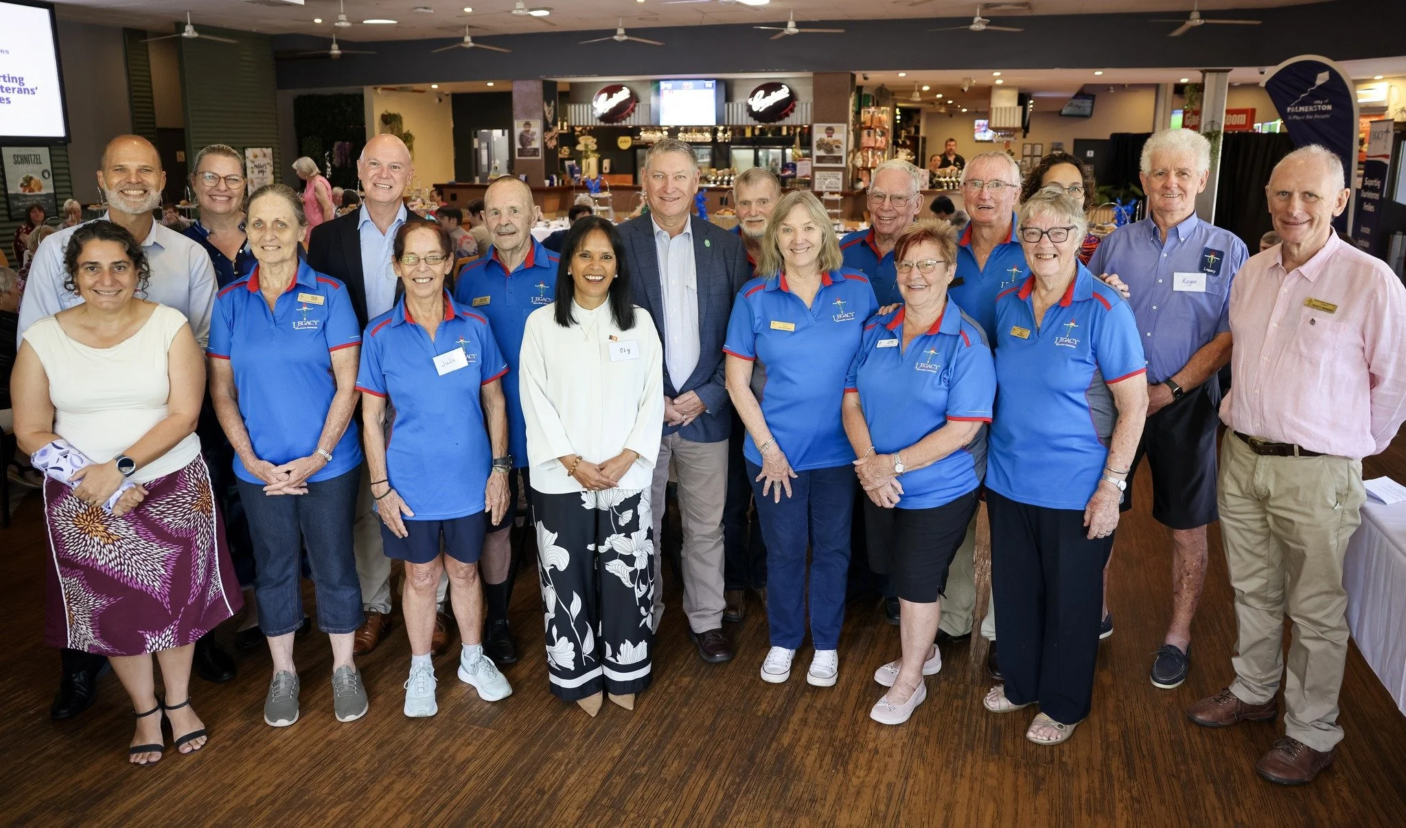 Group of volunteers and staff members posing together at an indoor event, some wearing blue uniforms with 'Legacy' logos and name tags, smiling for the camera.