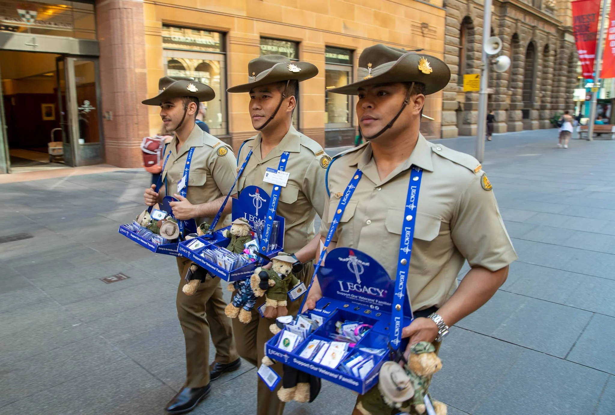 Three men in uniform walking on a city street, carrying boxes with items for a charity or fundraising event.