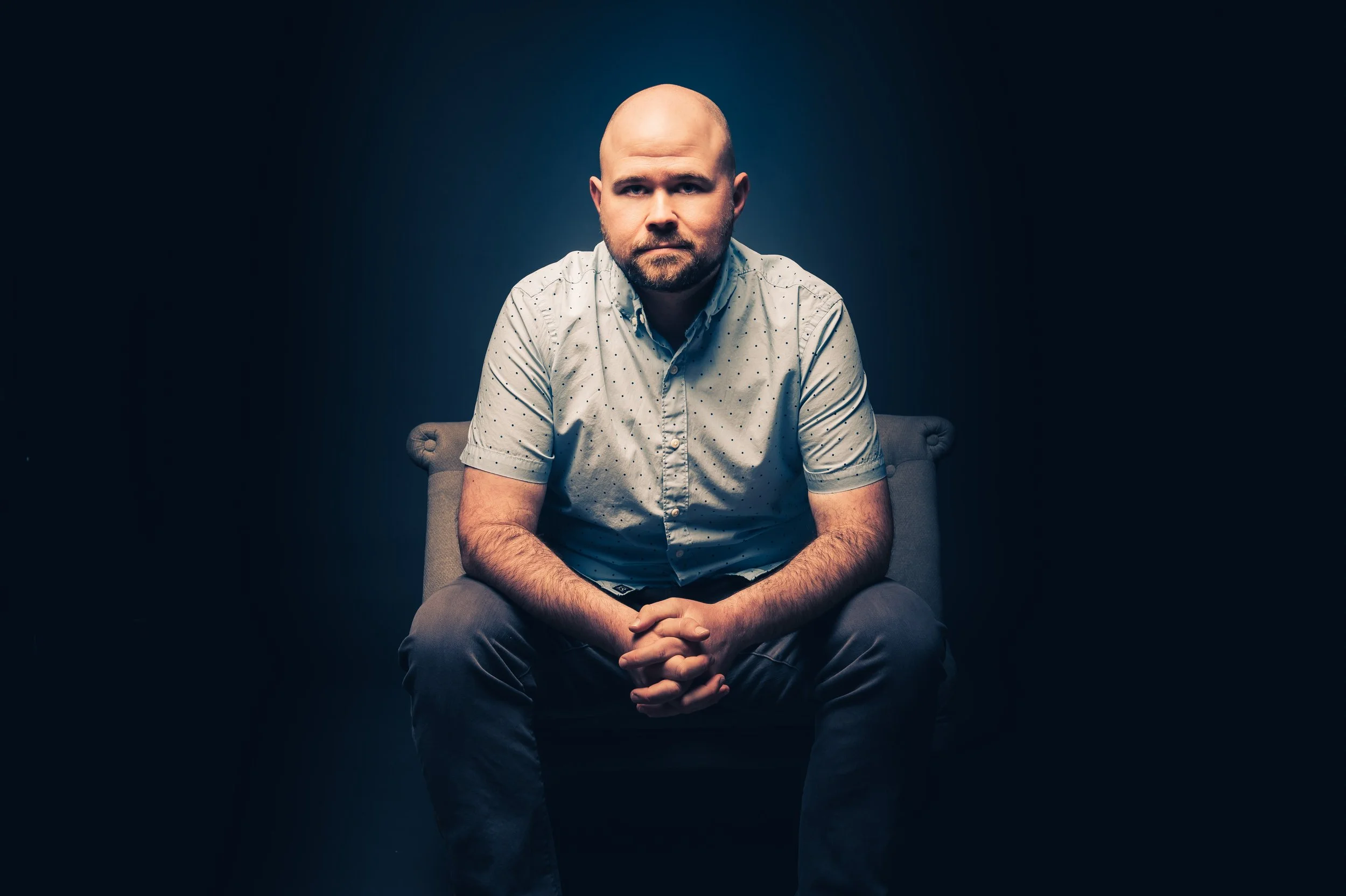 Bald man with beard sitting on a chair against a dark background, wearing a light-colored, polka-dotted shirt.