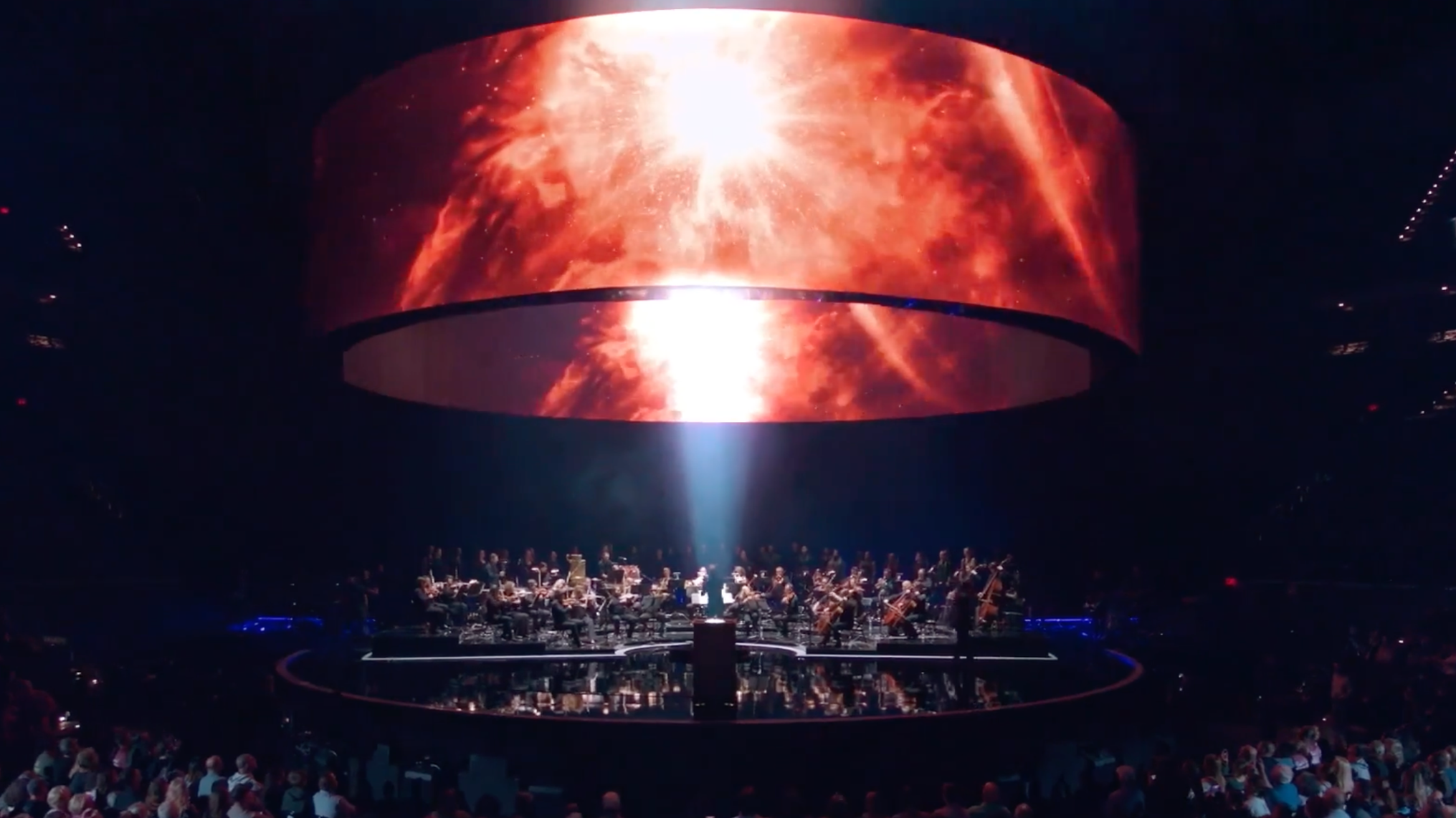 Orchestra performing in a dark concert hall with a large, curved screen displaying a fiery, cosmic explosion above them.
