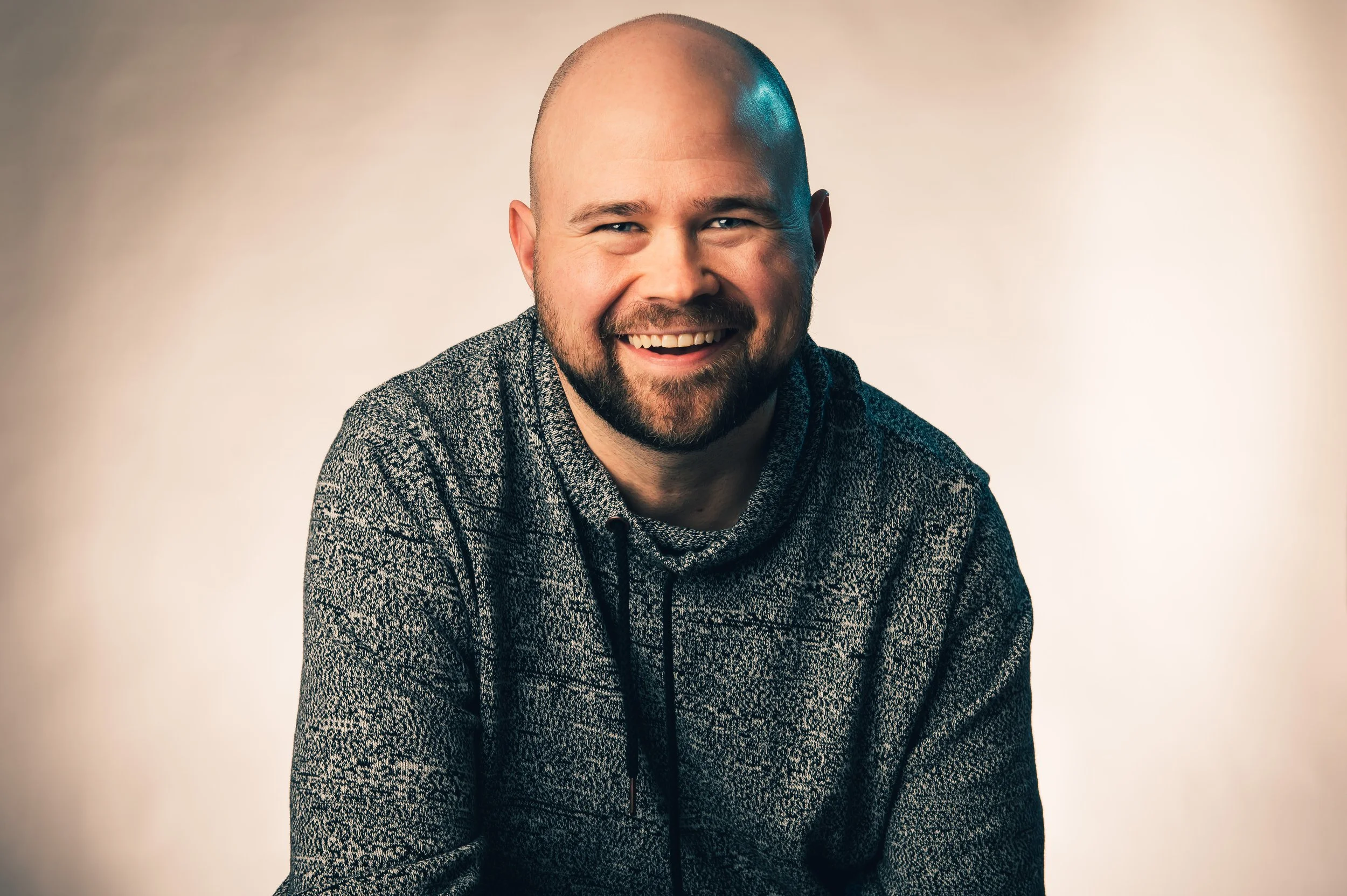 A smiling man with a shaved head and beard, wearing a patterned sweater, posing against a plain background.