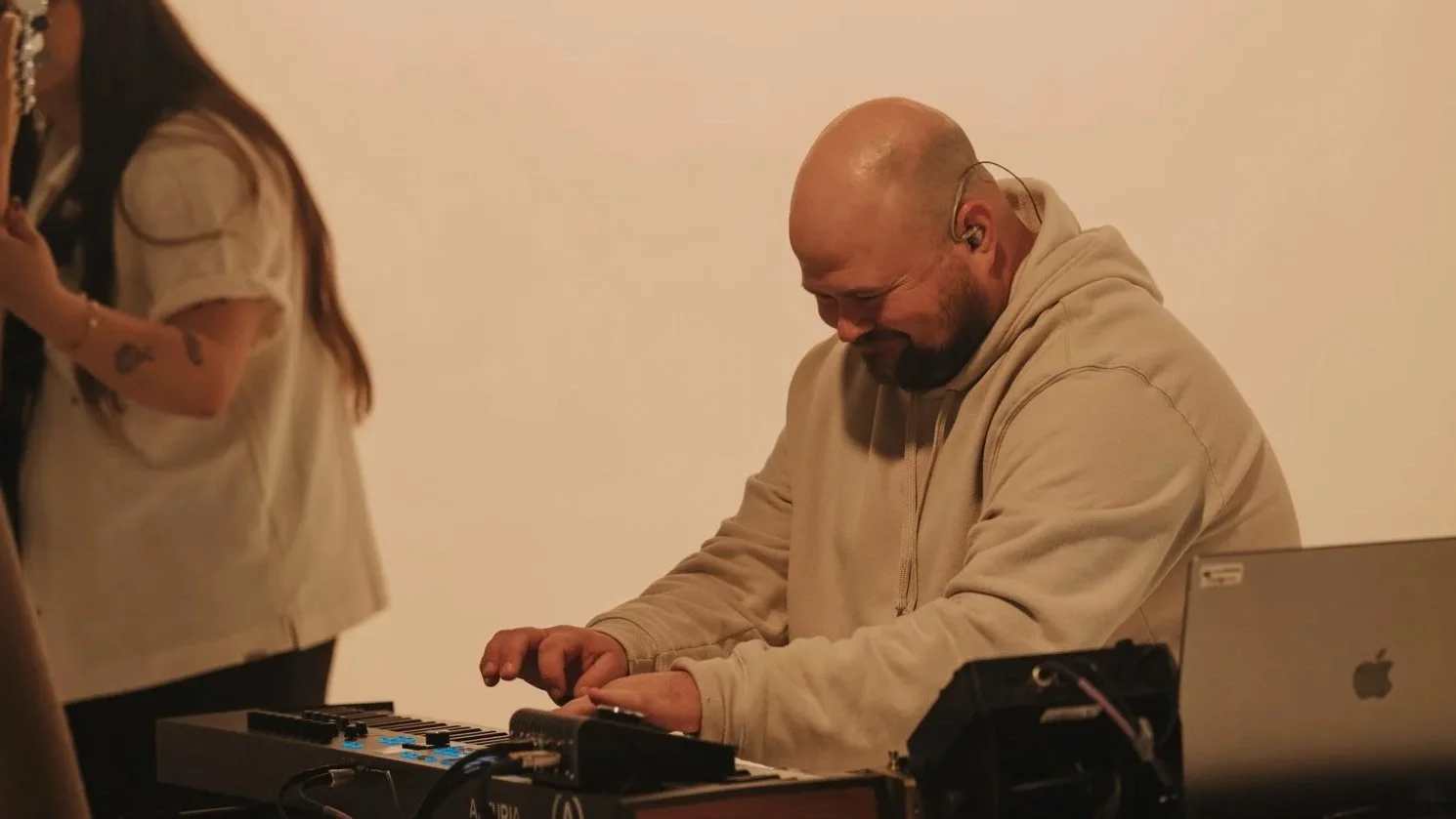 A man with a shaved head and beard, wearing a beige hoodie, smiling while playing a keyboard, with a laptop and audio equipment around him.