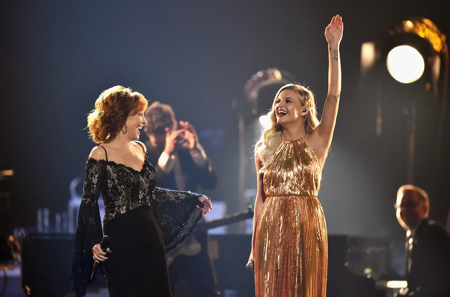 Two women on stage, one in a black lace dress holding a microphone, the other in a shiny gold dress, smiling and raising her hand, with stage lights in the background.