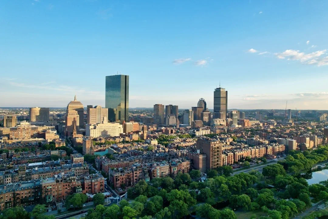 Aerial view of Boston skyline with tall buildings, green trees, and a river in the foreground on a clear day.