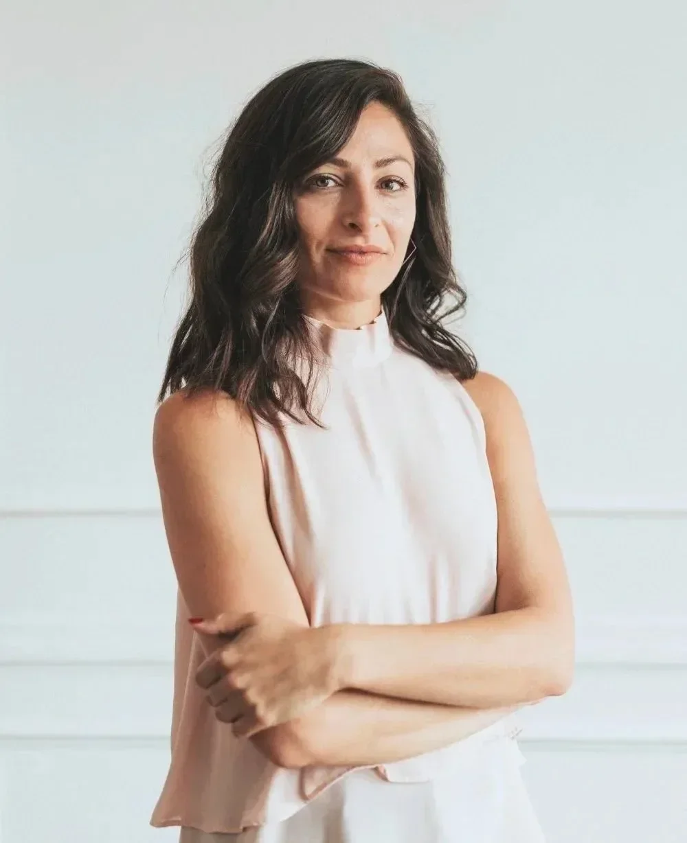 A woman with wavy dark hair wearing a sleeveless light pink top with her arms crossed, standing against a plain light background.