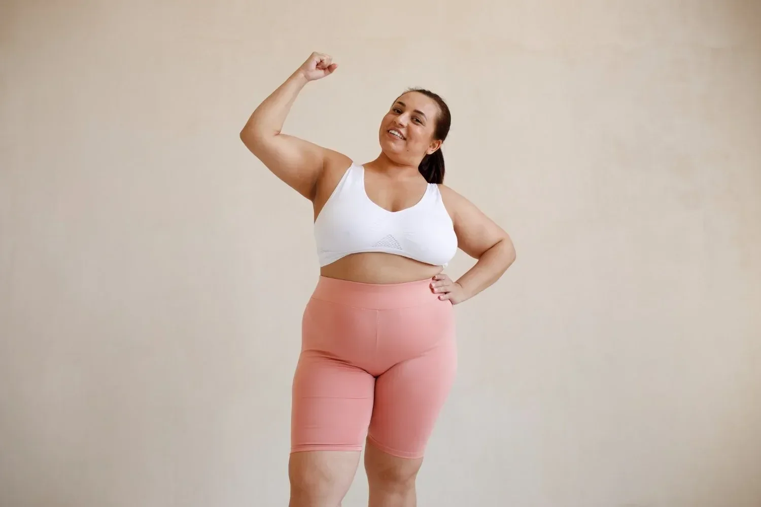 Plus-sized woman with dark hair tied back, smiling, wearing a white sports bra and peach-colored shorts, flexing her right arm in a victory pose over a plain beige background.