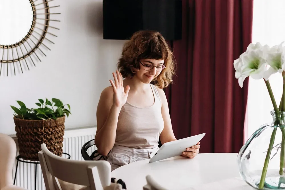 Smiling young woman with curly hair and glasses waving while looking at a tablet at a dining table with a vase of white flowers and a potted plant in the background.