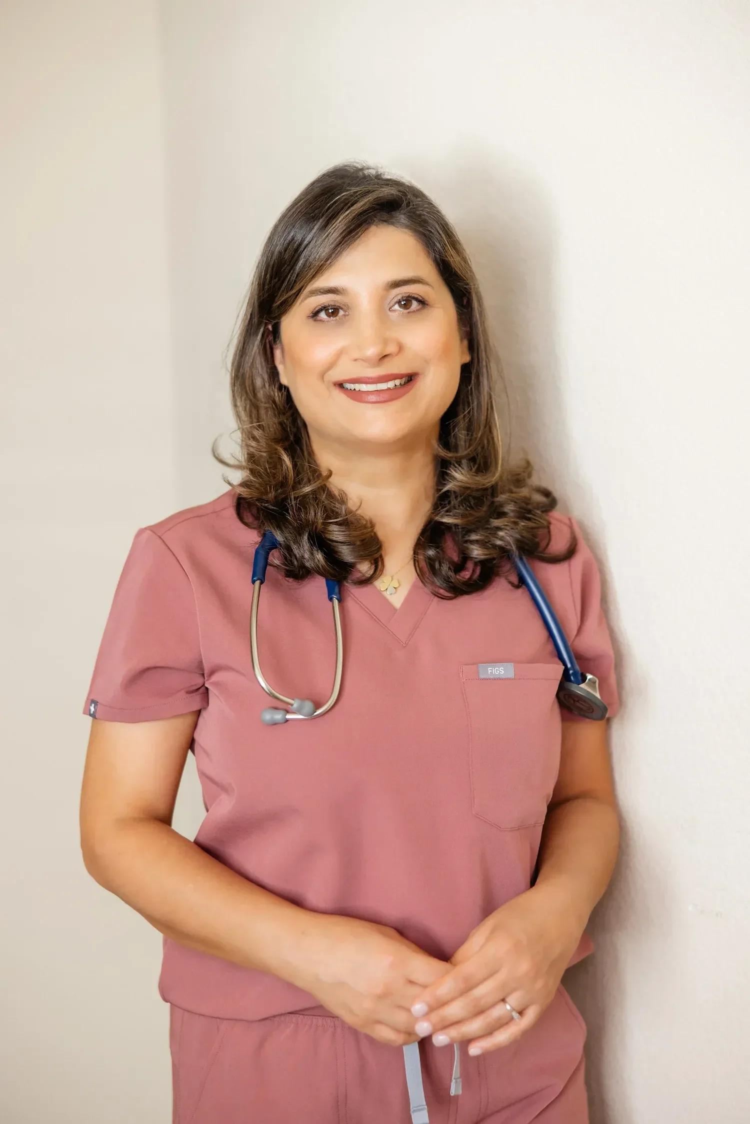Dr. Sima Kavand wearing pink medical scrubs with a stethoscope around her neck, standing against a white wall, smiling at the camera.