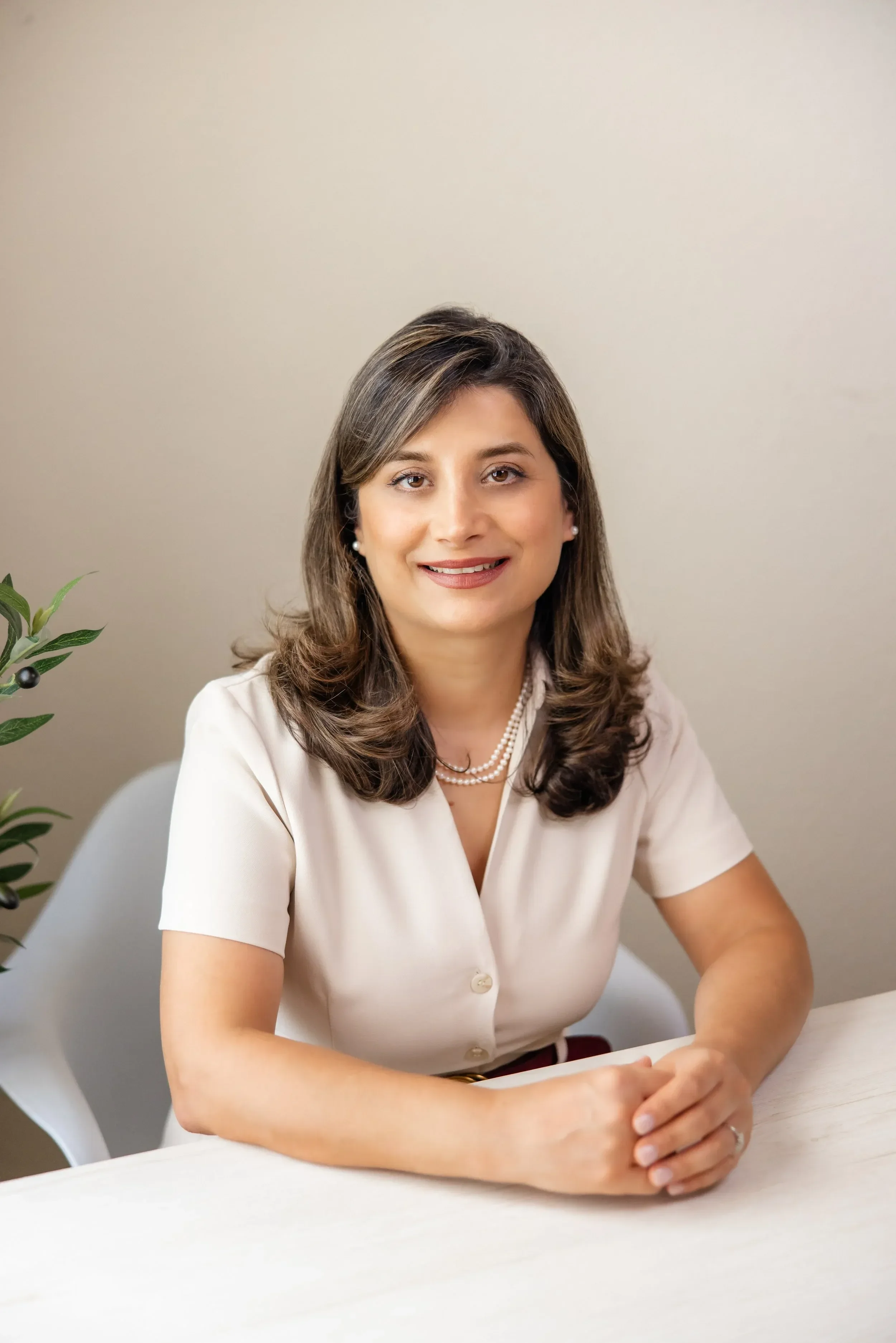 Dr. Sima Kavand wearing a white short-sleeved blouse and a pearl necklace, with her hands folded on the desk. The background is a plain beige wall, with a small green plant visible on the left side of the image.