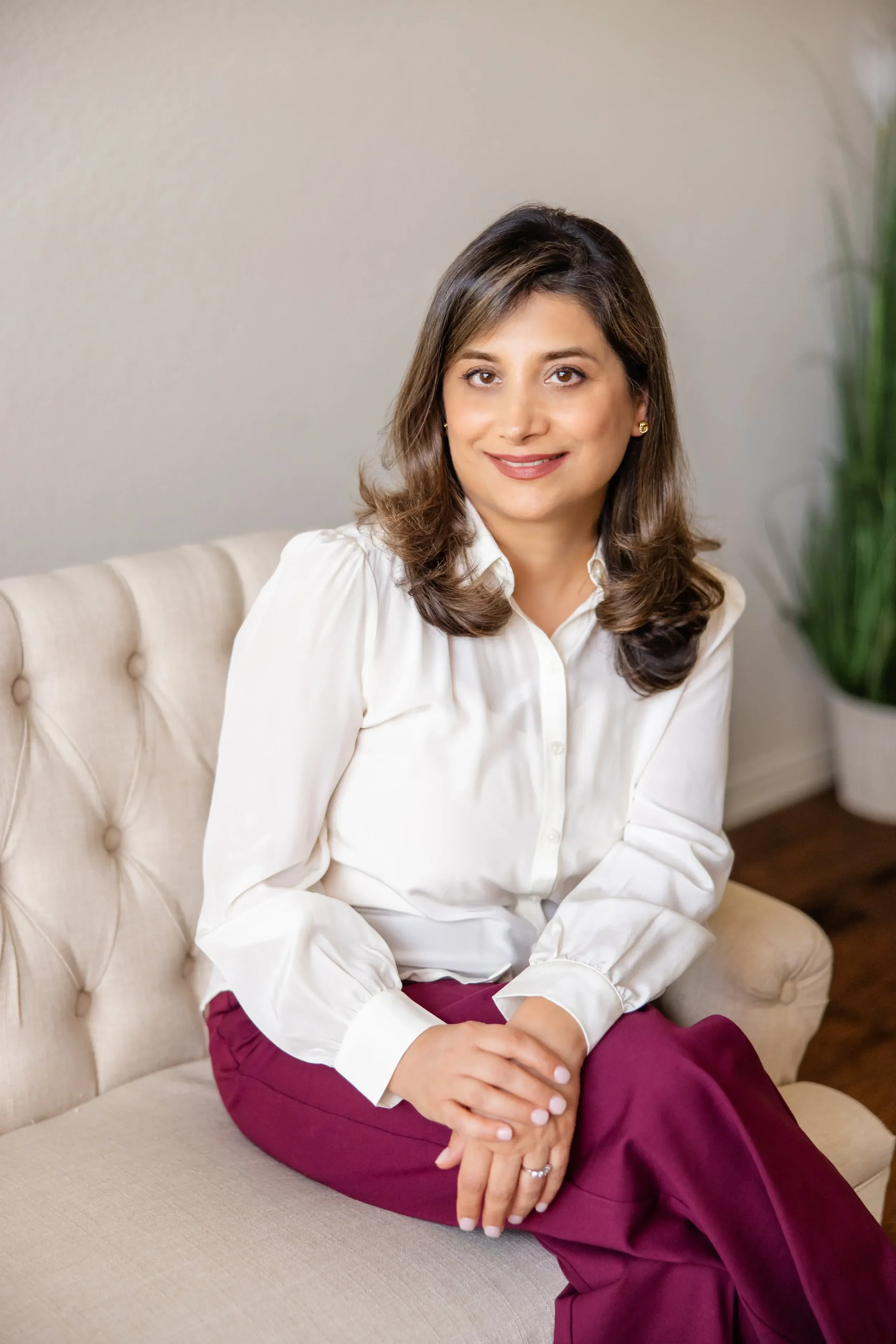 Dr. Sima Kavand sitting on a beige sofa, wearing a white shirt and maroon pants, with dark brown hair, smiling, and holding her hands on her lap. There is a green plant partially visible on the right side of the image.