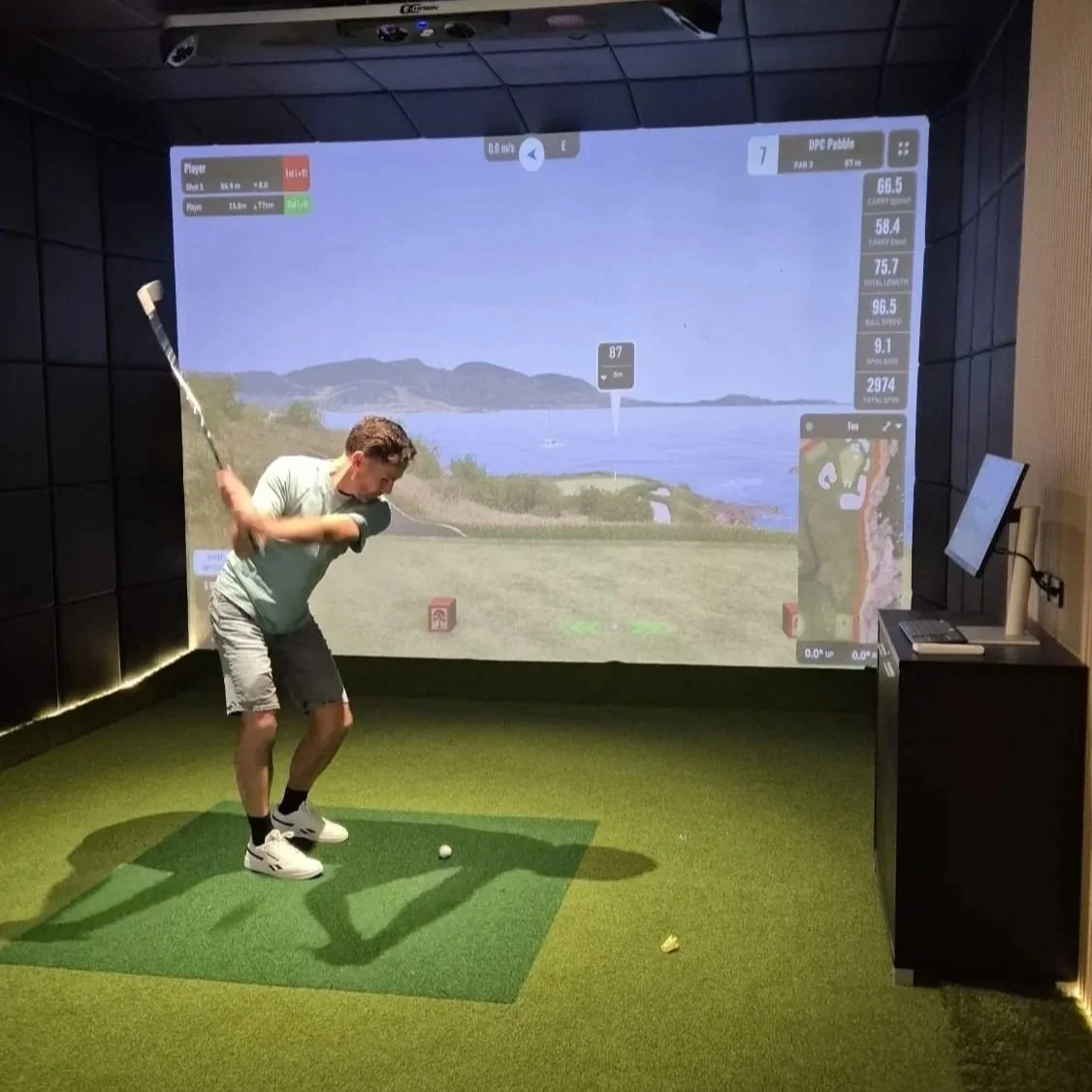 Man practicing golf swings in an indoor golf simulator, with a scenic view of water and mountains projected on the wall.