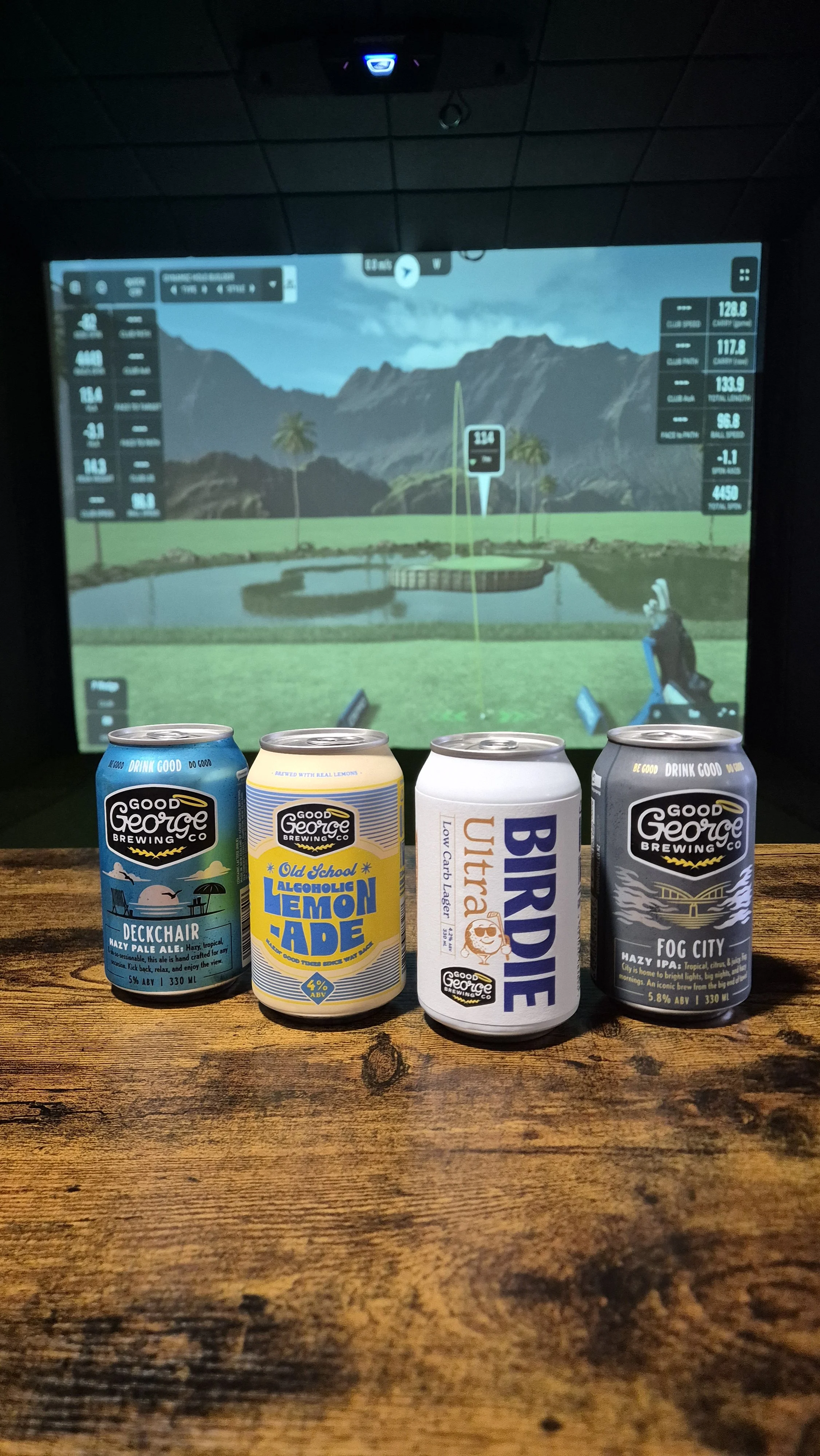 Four cans of craft beer on a wooden table with a golf simulation screen in the background