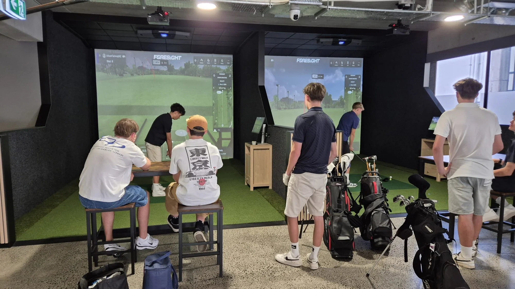Several people inside a golf simulation room with large screens displaying virtual golf courses. Some are standing, others sitting, with golf bags and clubs nearby.
