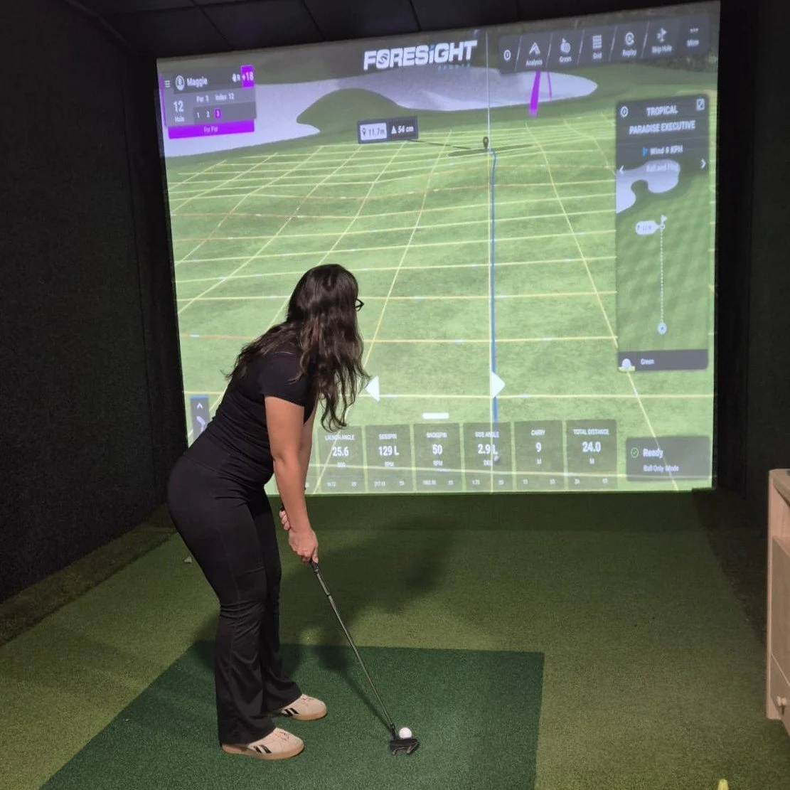 A woman in black pants and a black shirt preparing to hit a golf ball in an indoor golf simulator, with a large electronic screen displaying a virtual golf course and shot data.