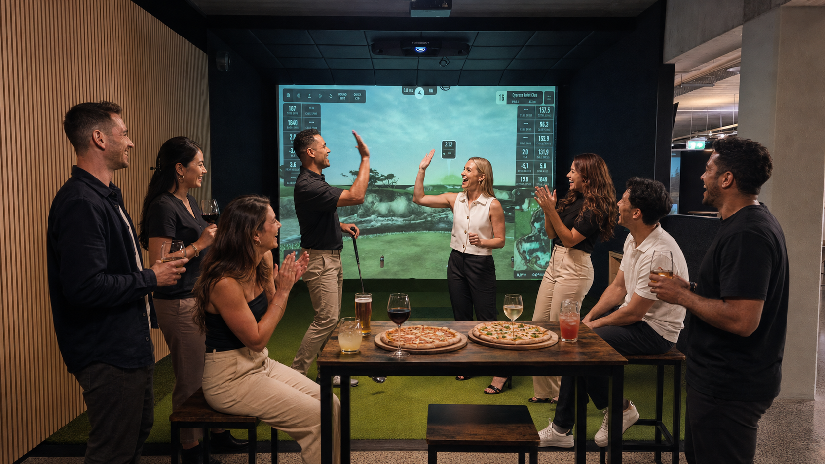 Group of people celebrating and high-fiving in front of a golf simulation screen in a bar with pizza and drinks on the table.