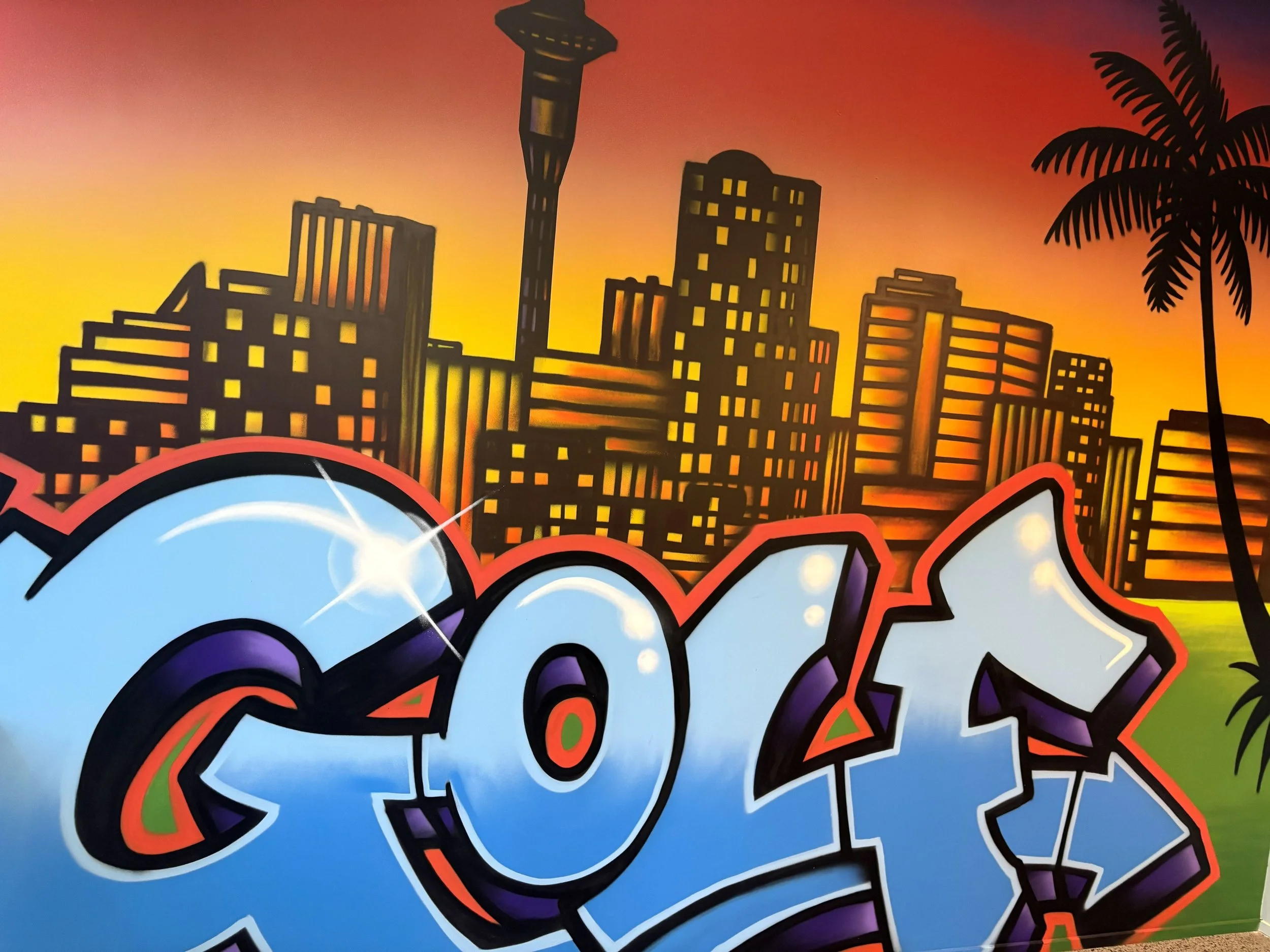 Graffiti-style art depicting a city skyline silhouette with buildings and a tall tower, set against a colorful sunset background with orange and purple hues, and a palm tree on the right side.